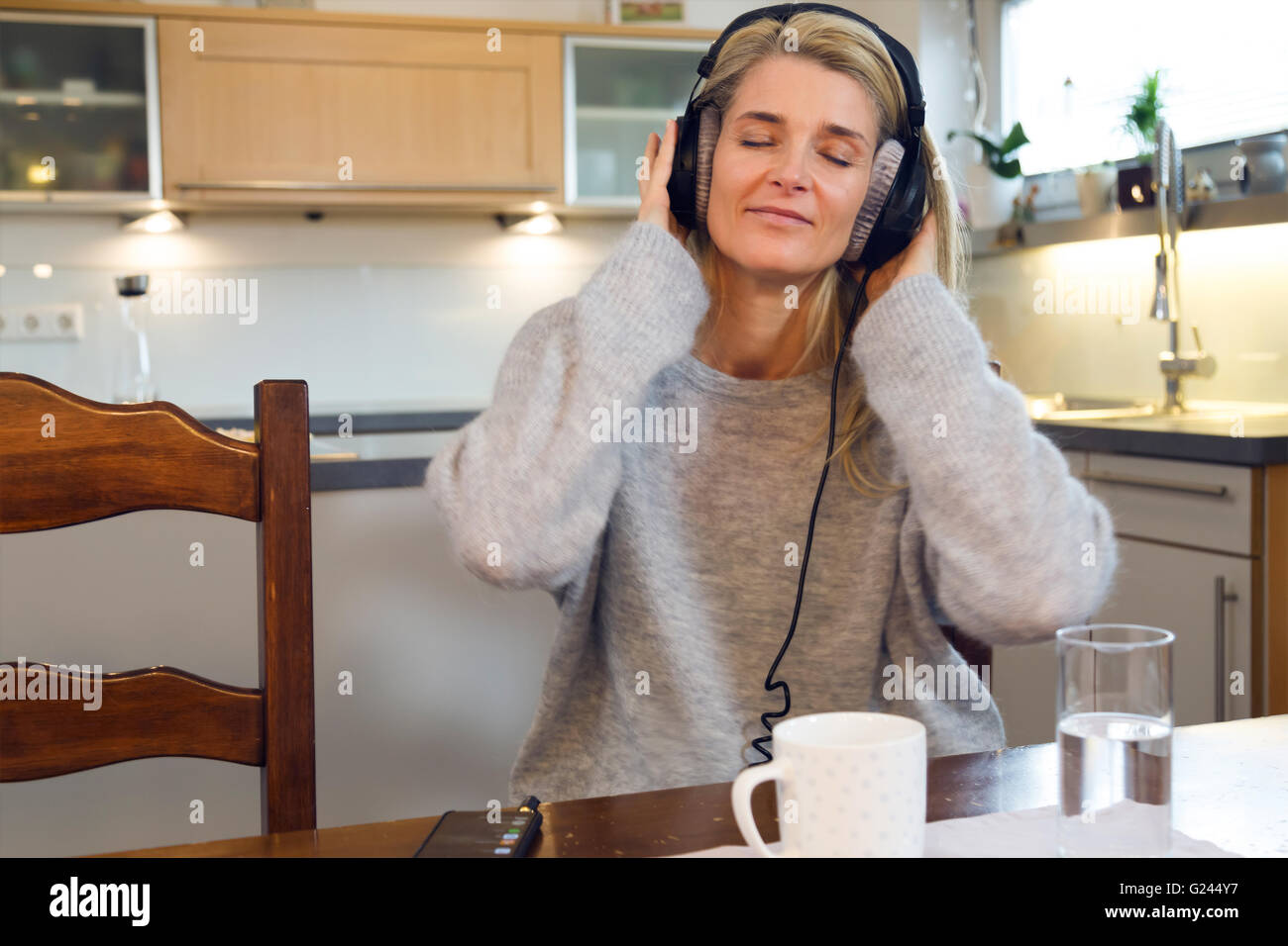 Frau mittleren Alters genießen Sie Musik auf Ihrem Kopfhörer. Zu Hause, entspannt und wohl. Moderne Einrichtung hinter ihr. Sitzt. Mittellange Aufnahme. Stockfoto