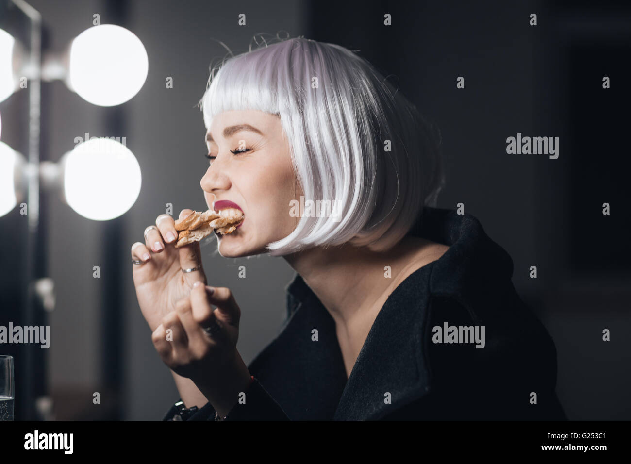 Lustige nette junge Frau in blonde Perücke Essen Pizza in Umkleidekabine Stockfoto