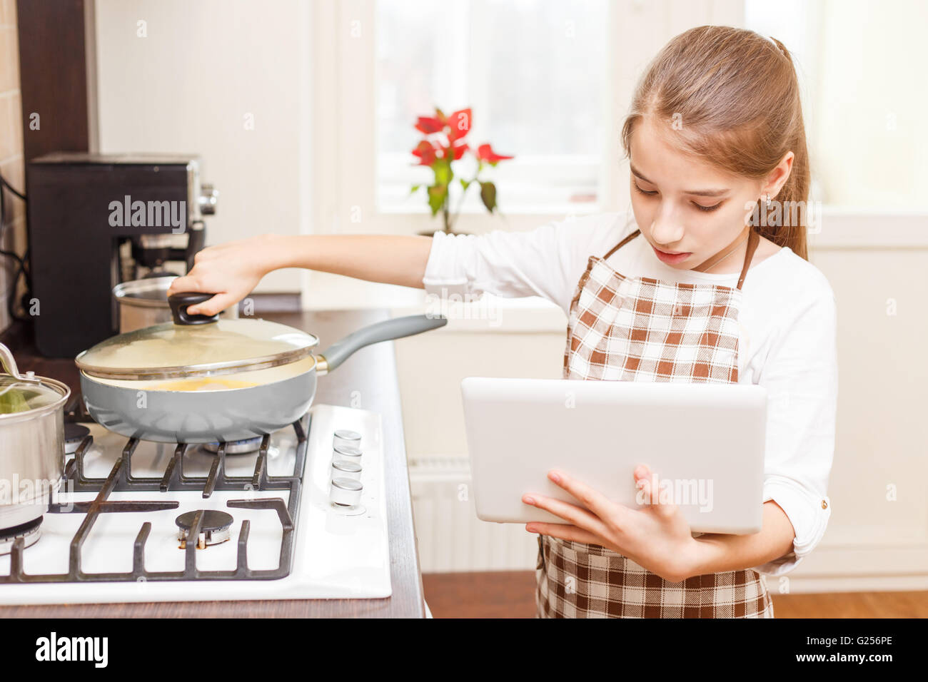 Kleinen Teenager-Mädchen Kochen am Herd mit Laptop. Teenager Mädchen lernen kochen online Stockfoto