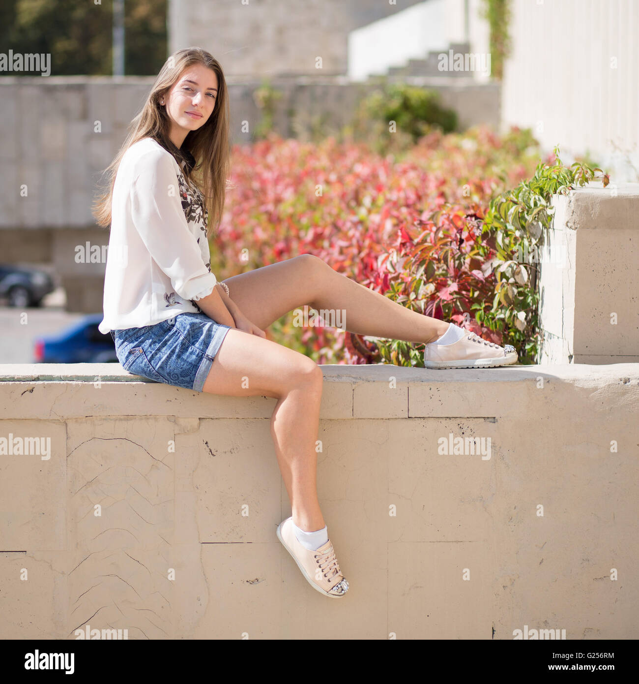 Junge Mode-Studentin auf der Mauer sitzen. Outdoor Portrait lächelnd Mädchen in kurzen Hosen und weißen Hemd Stockfoto