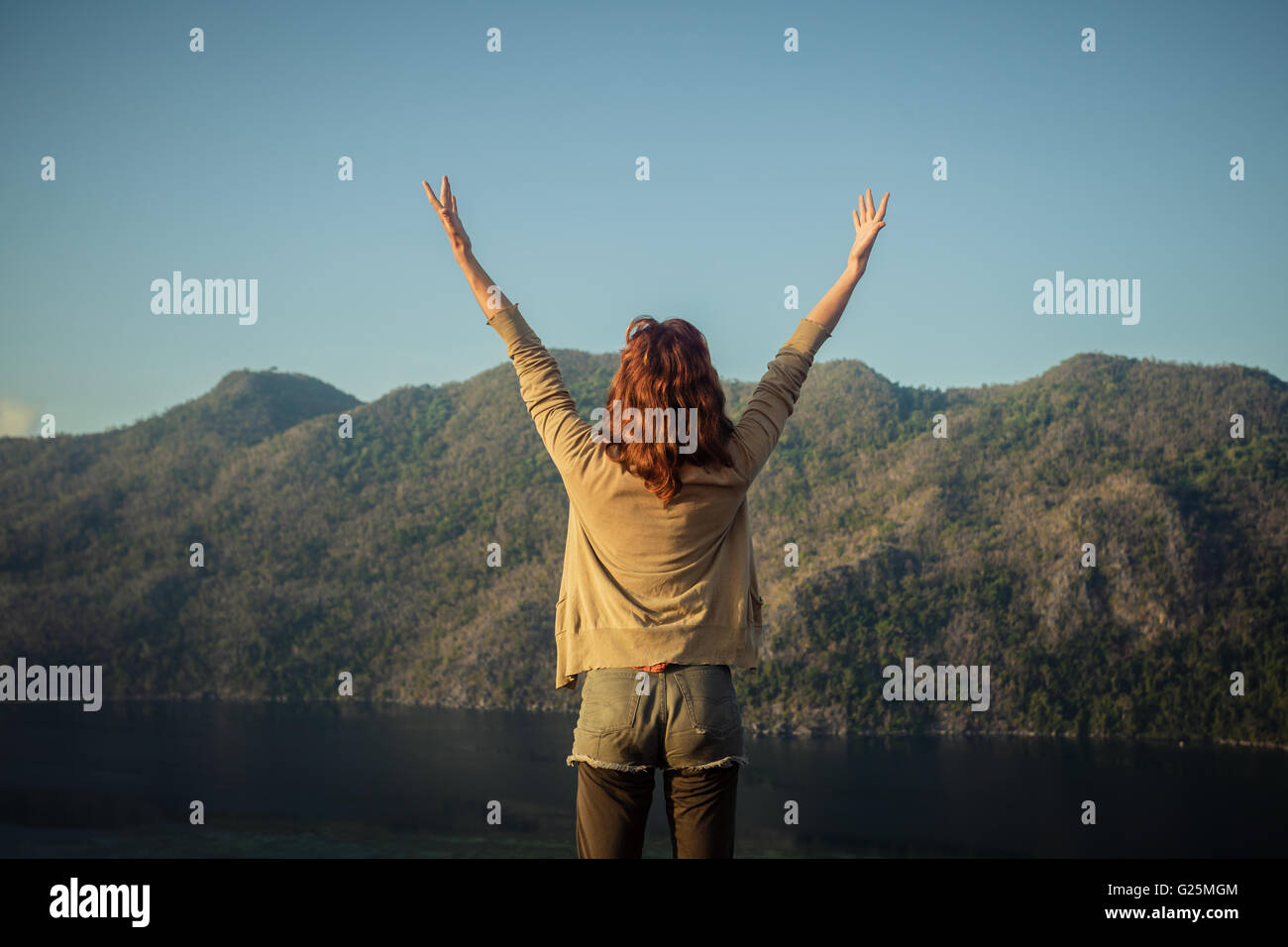 Eine glückliche junge Frau hebt ihre Arme auf einem Berg mit Blick auf eine Bucht in einem tropischen Land Stockfoto