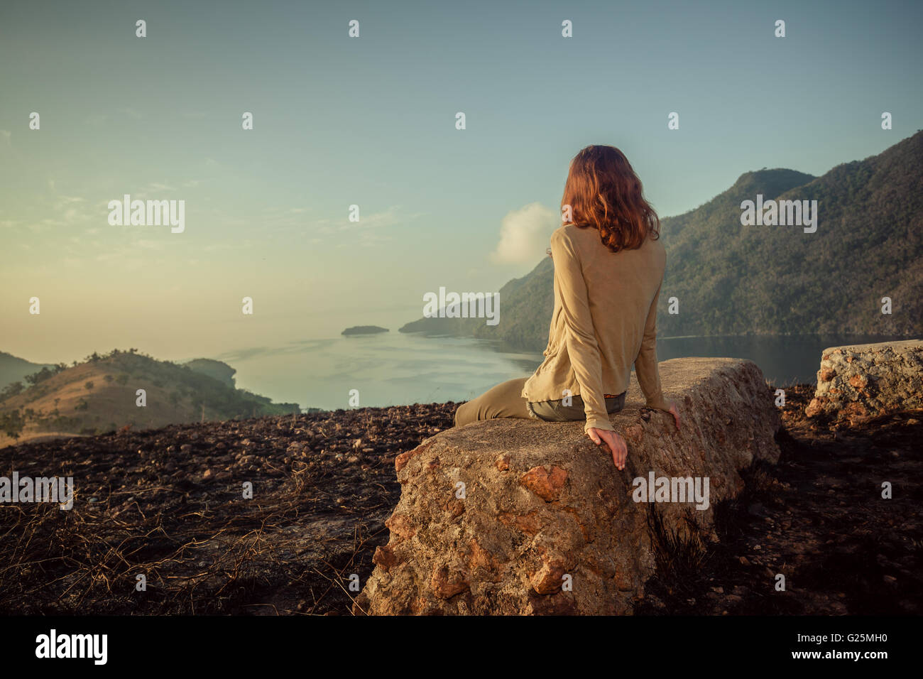 Eine junge Frau sitzt auf einem ungewöhnlichen Felsen auf einem Berg mit Blick auf eine Bucht bei Sonnenaufgang in einem tropischen Klima Stockfoto