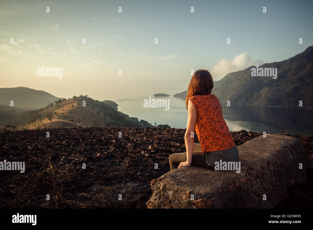 Eine junge Frau sitzt auf einem ungewöhnlichen Felsen auf einem Berg mit Blick auf eine Bucht bei Sonnenaufgang in einem tropischen Klima Stockfoto