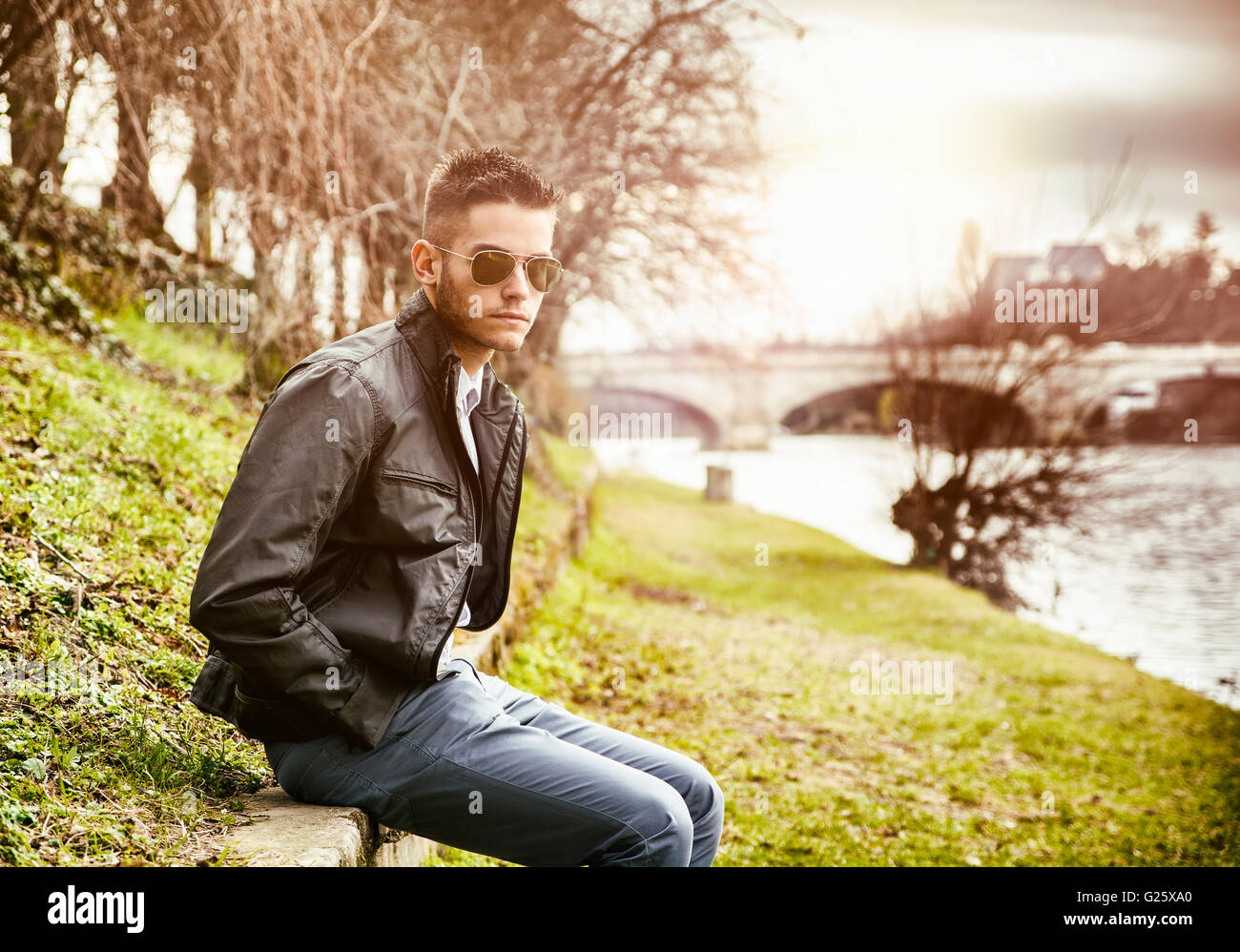 3/4 Länge des kontemplativen leichte Braunhaarige junge Mann trägt graue Jacke und Denim Jeans sitzen an Wand neben pi Stockfoto