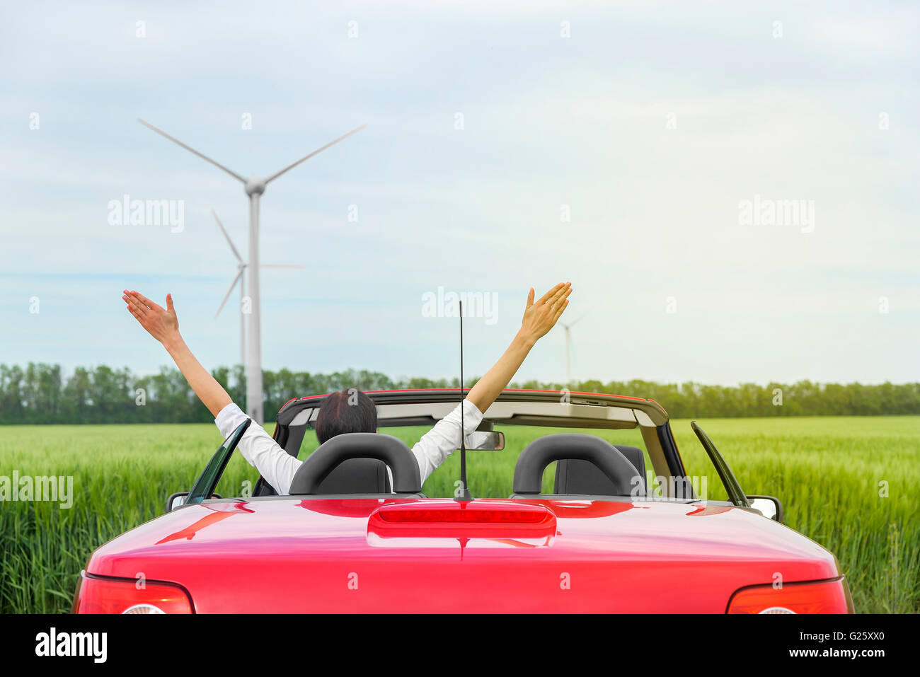 Frau in einem roten Cabrio in einem Feld mit Windkraft, erhebt Arme. Reisekonzept. Stockfoto