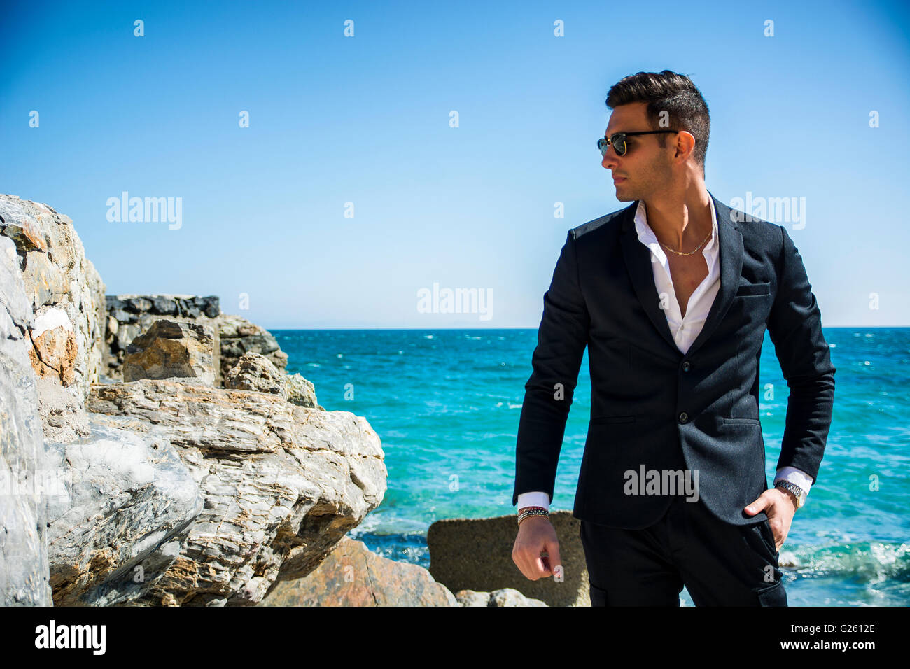 Schönen jungen Mann im klassischen Anzug am Strand halten Sonnenbrille beim wegschauen. Wellen des Meeres im Hintergrund Stockfoto