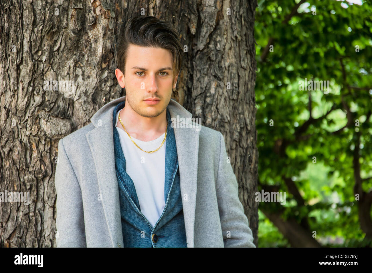 Hübscher junger Mann lehnte sich gegen Baum, Blick in die Kamera, an einem sonnigen Tag trägt einen leichten grauen Mantel Stockfoto