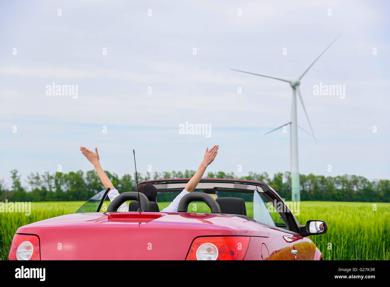 Frau in einem roten Cabrio in einem Feld mit Windkraft, erhebt Arme. Reisekonzept. Stockfoto