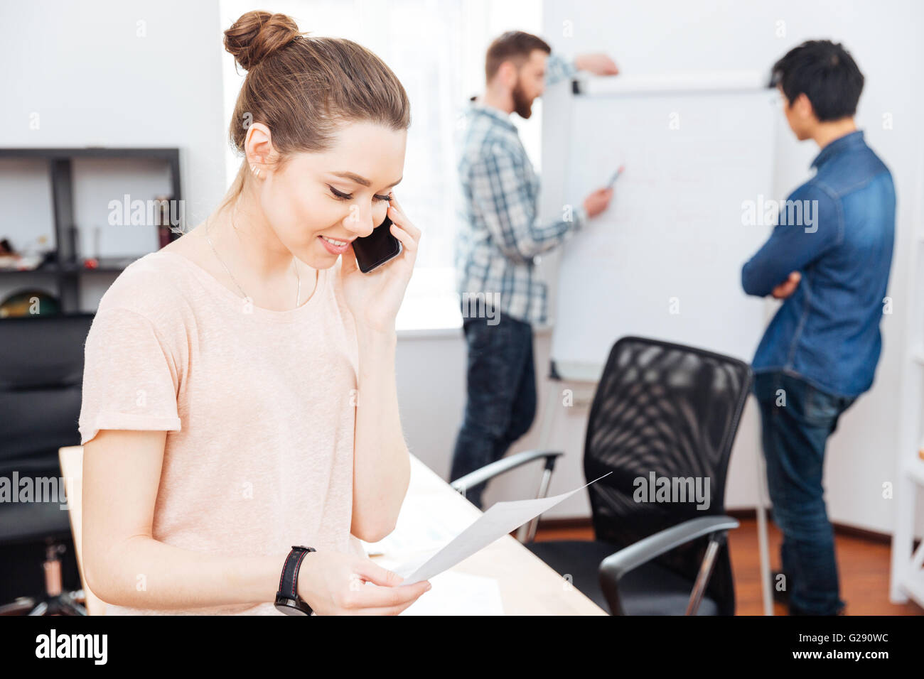 Fröhliche junge Geschäftsfrau telefonieren mit Handy während ihrer Kollegen arbeiten mit Flipchart in office Stockfoto