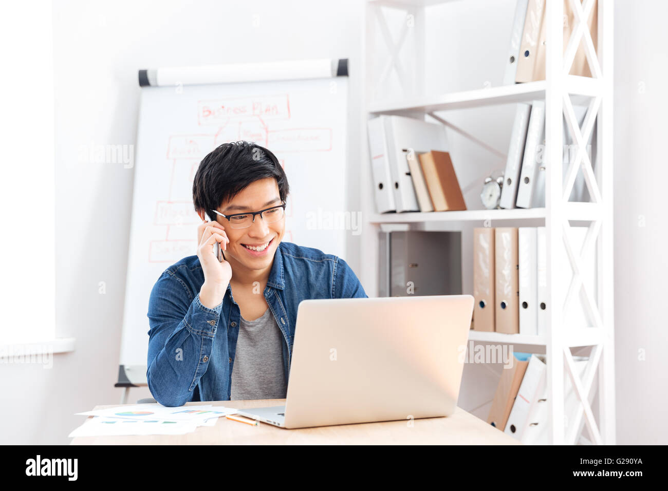 Glücklich hübscher junger Geschäftsmann in Gläsern mit Laptop und Handy telefonieren im Büro Stockfoto