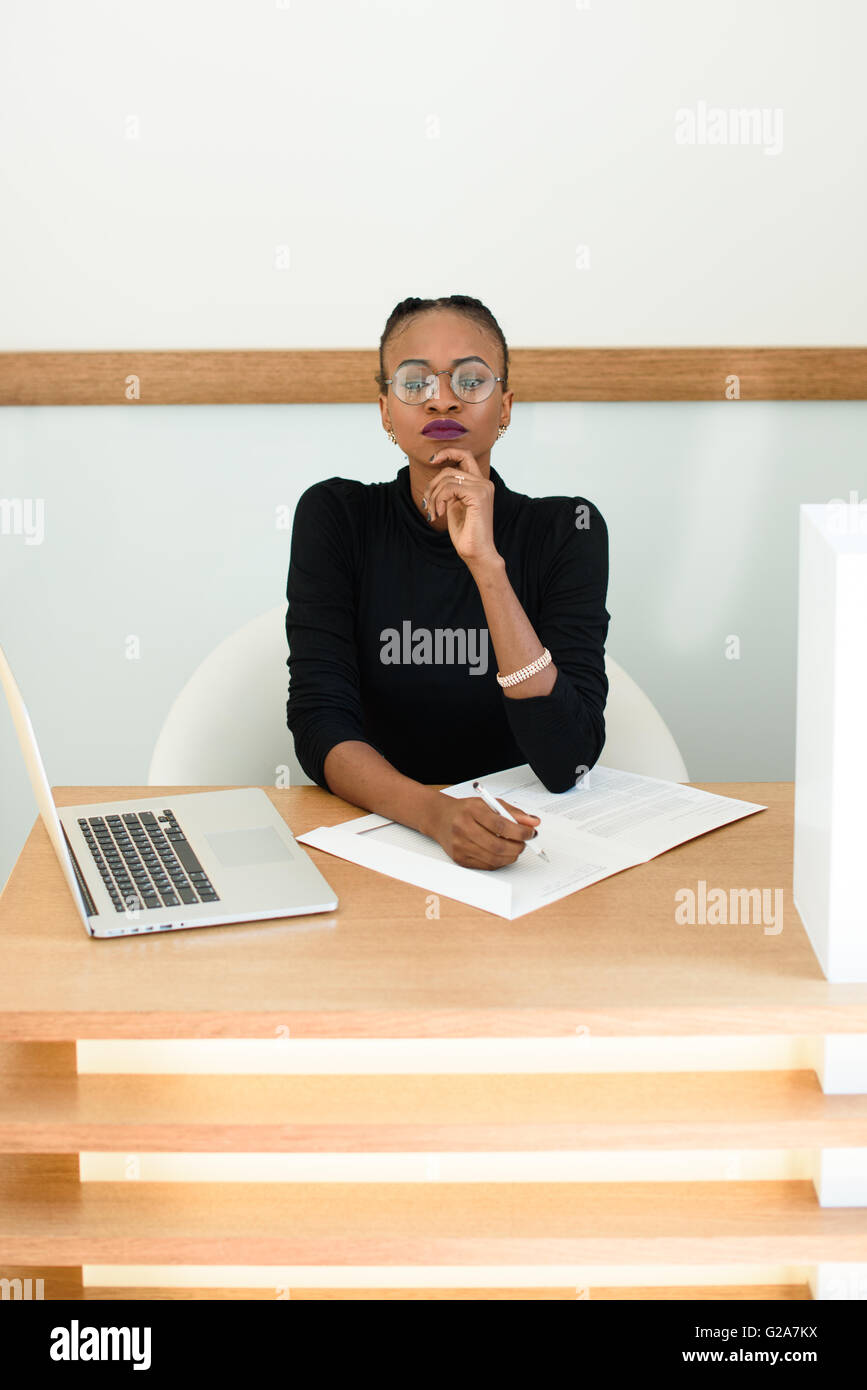 Geschäftsfrau mit Brille, Blick in die Kamera halten Kinn mit Notizblock und Laptop am Schreibtisch im Büro arbeiten Stockfoto
