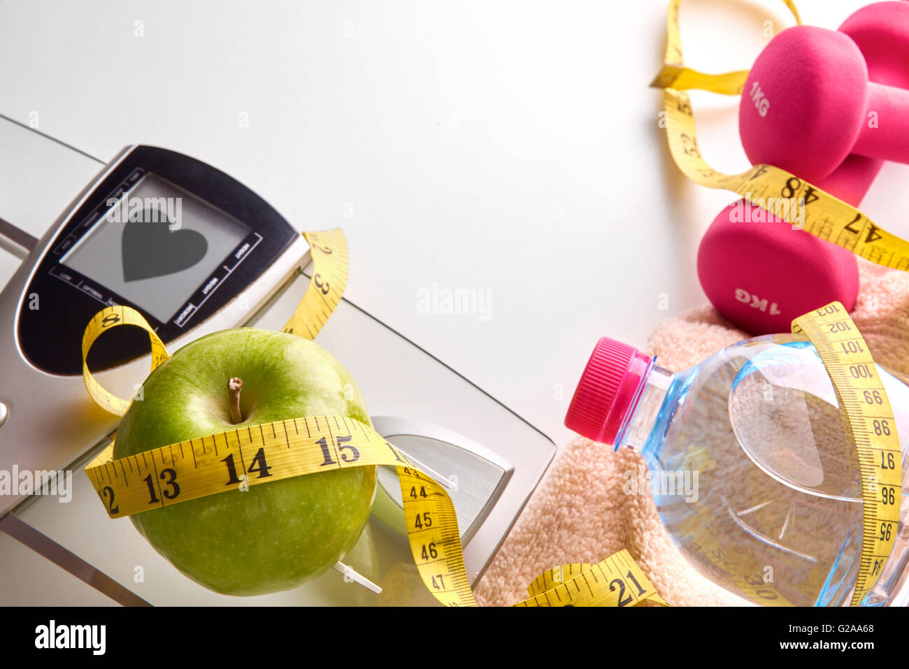 Skalieren Sie mit gesunden Herzen Nachricht, rosa Hanteln, Apfel, Flasche Mineralwasser, Handtuch und Maßband auf weißen Tisch. Stockfoto