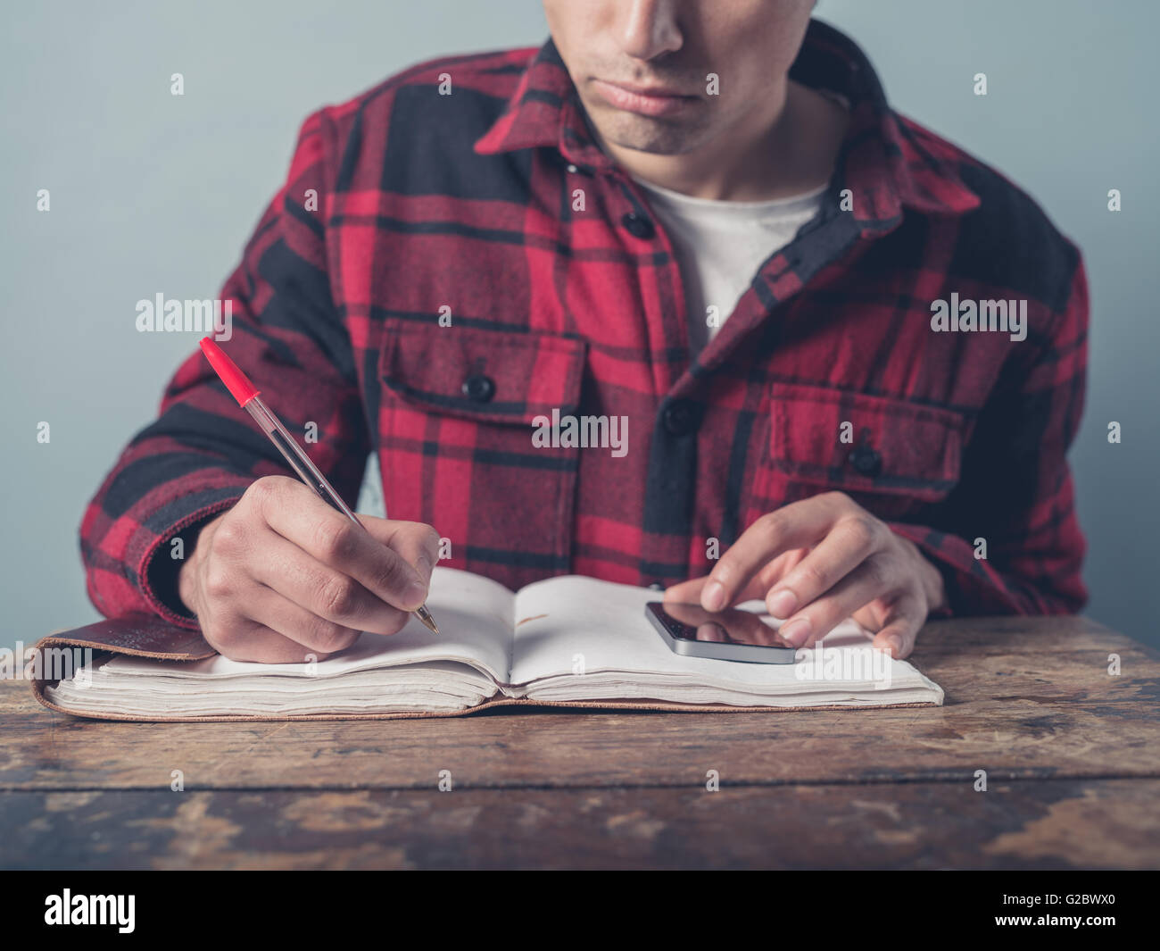 Ein junger Mann mit einer karierten Jacke ist per Smartphone und ist Notizen in einem Notizbuch an einem Schreibtisch Stockfoto