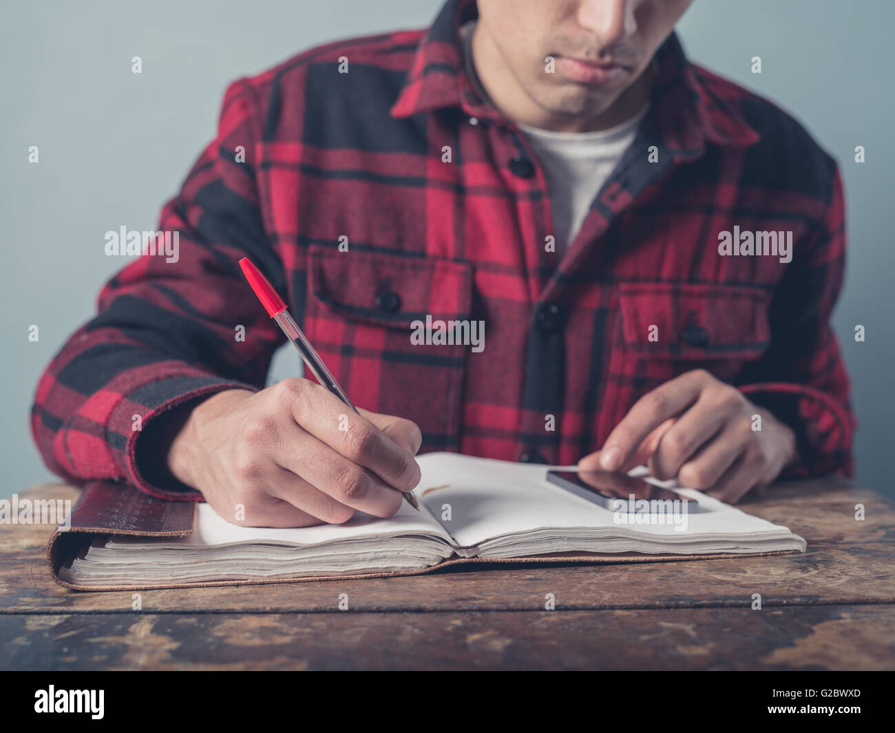 Ein junger Mann mit einer karierten Jacke ist per Smartphone und ist Notizen in einem Notizbuch an einem Schreibtisch Stockfoto