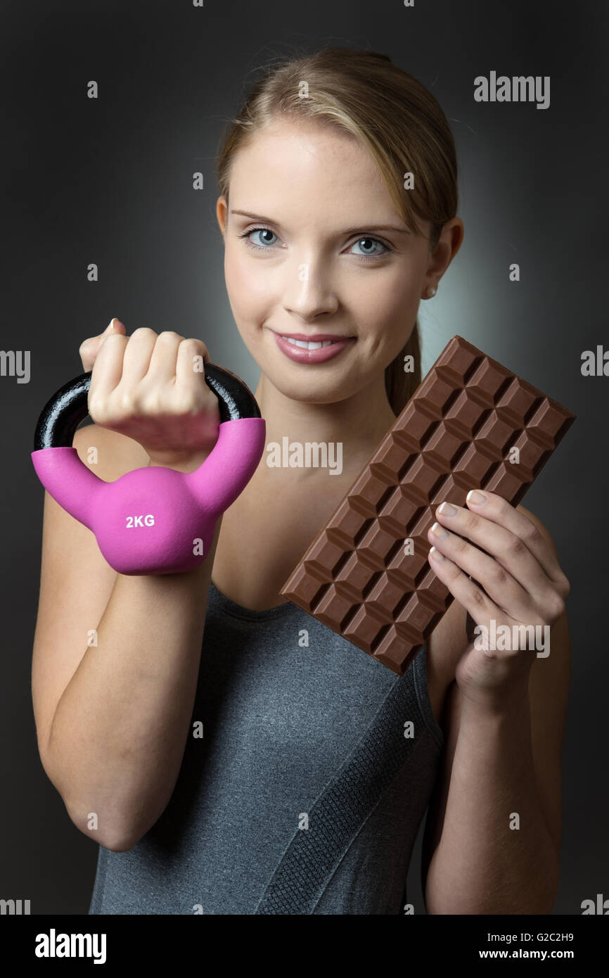 Nahaufnahme Bild von einer hübschen jungen Fitness-Modell hält eine rosa Wasserkocher Glocke und eine große Tafel Schokolade, erschossen auf einem grauen Hintergrund Stockfoto
