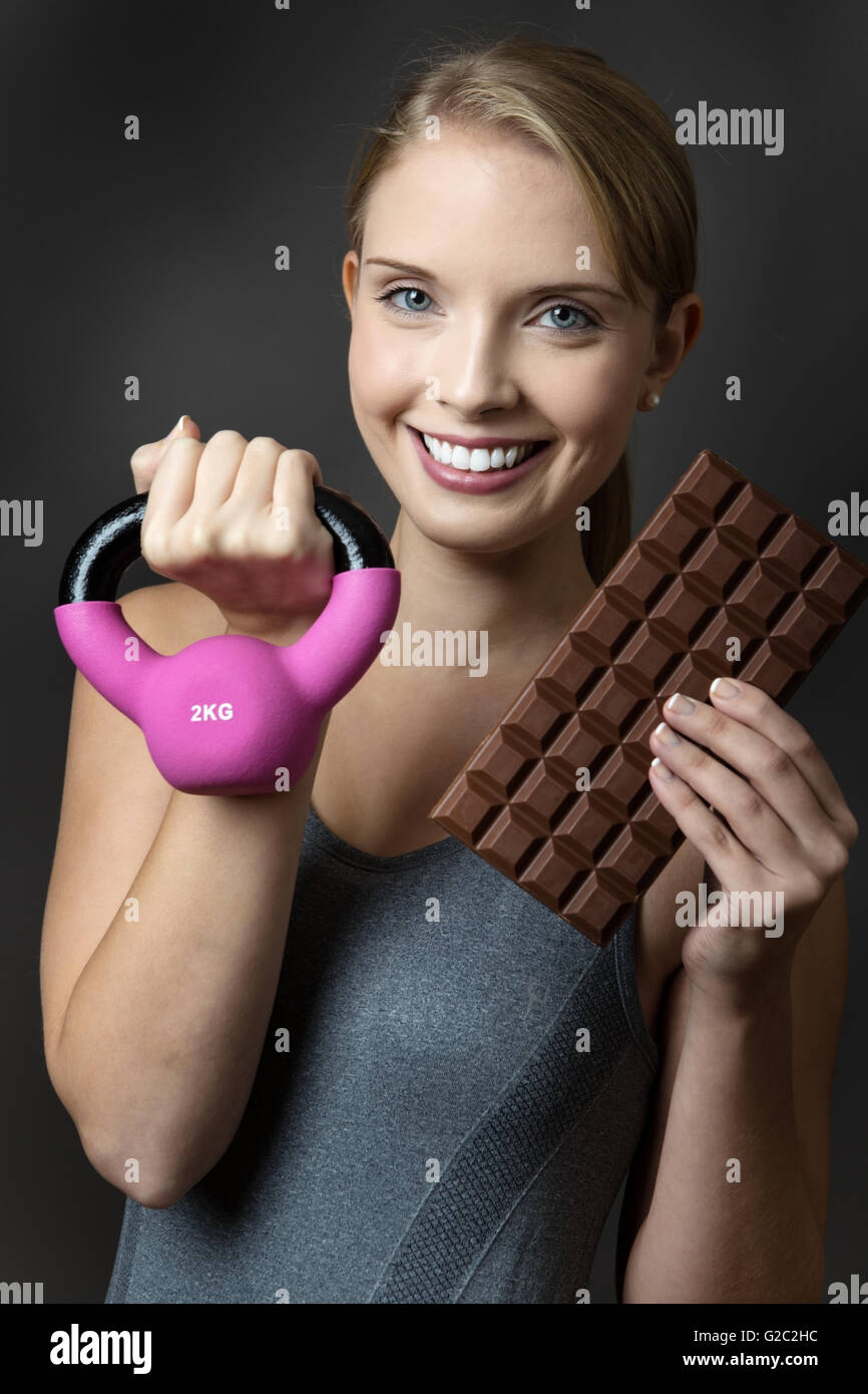 Nahaufnahme Bild von einer hübschen jungen Fitness-Modell hält eine rosa Wasserkocher Glocke und eine große Tafel Schokolade, erschossen auf einem grauen Hintergrund Stockfoto