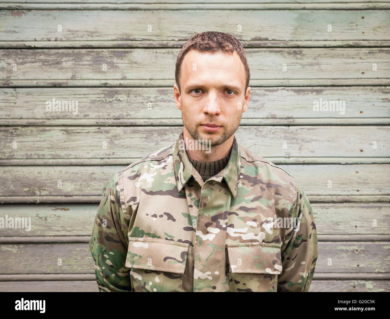 Ernsthafte kaukasischen Jüngling in militärischen Tarnuniform. Outdoor Closeup Portrait über grüne ländliche Holzwand Stockfoto