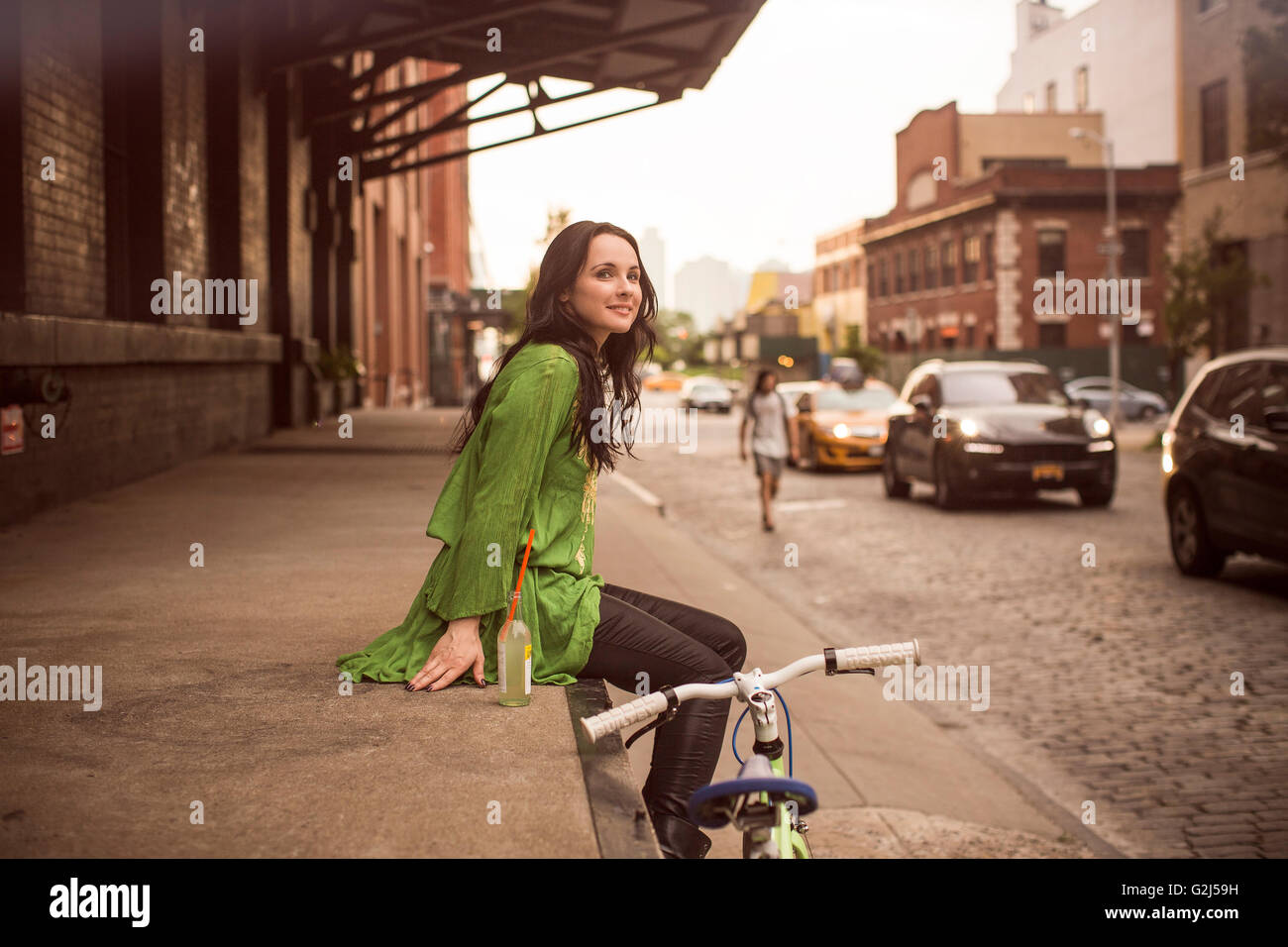 Frau mit Getränk sitzen beim Laden der Plattform neben Fahrrad auf gepflasterten Straße, New York City, USA Stockfoto