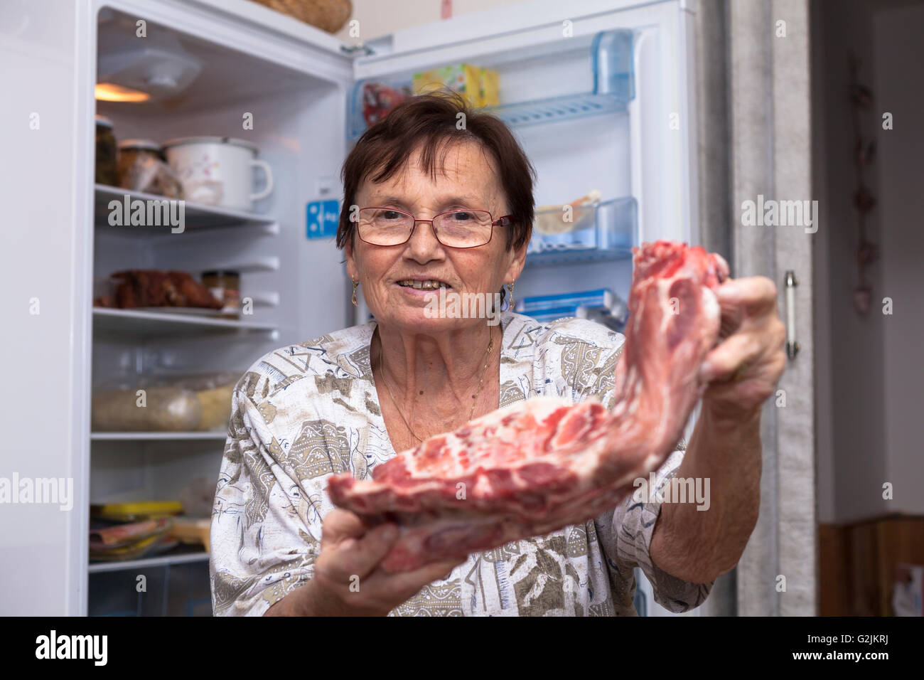 Glücklich senior Frau hält rohe Schweinerippchen vor den offenen Kühlschrank steht in der Küche. Stockfoto