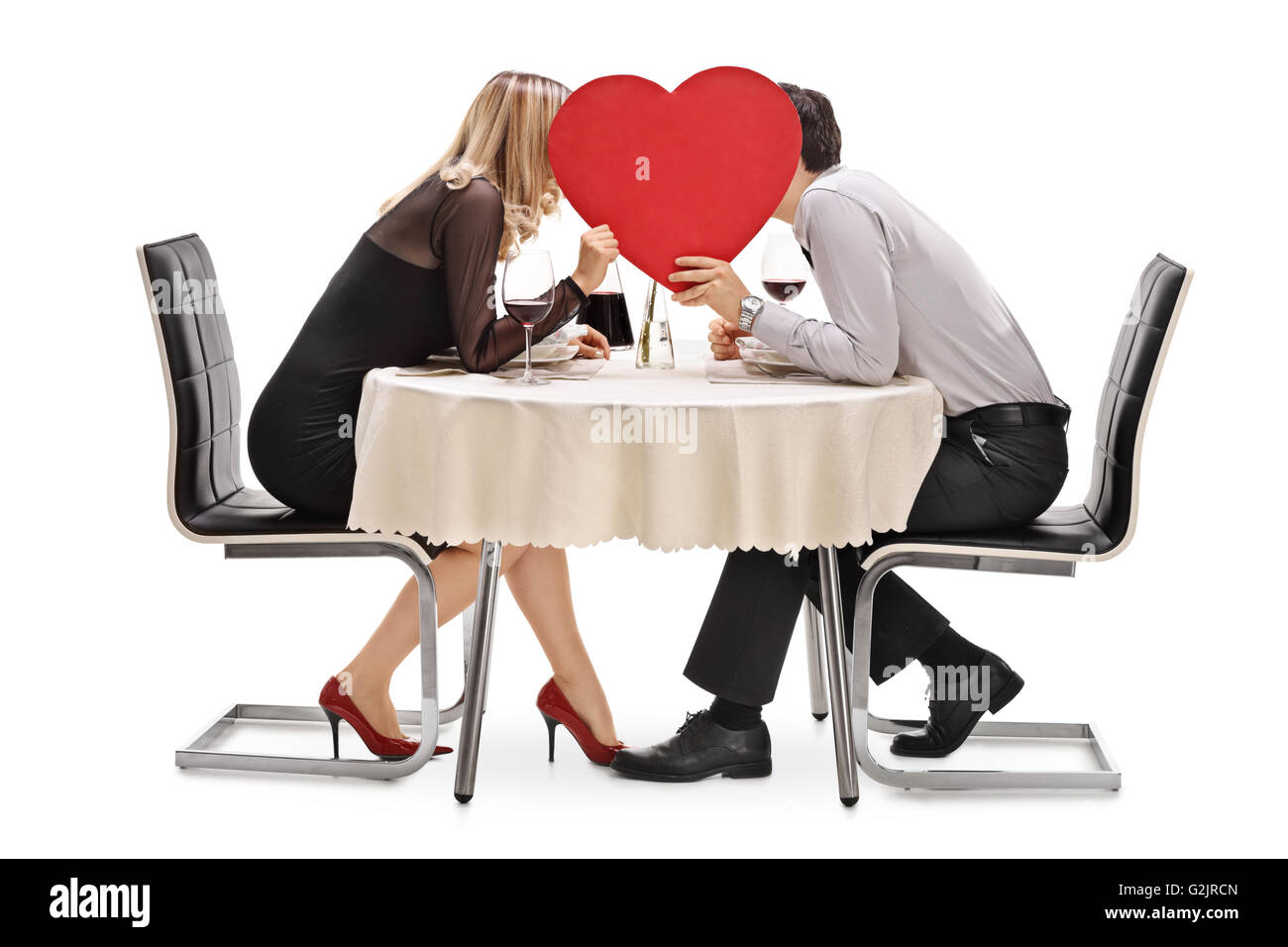 Junge Paar küssen hinter einem großen roten Herz sitzt auf einem Tisch im Restaurant isoliert auf weißem Hintergrund Stockfoto