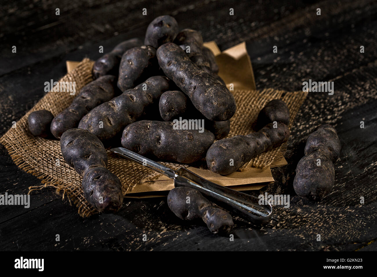 Blaue, violette Kartoffeln und Schäler auf braunem Papier, Jute und dunkles Holz Stockfoto