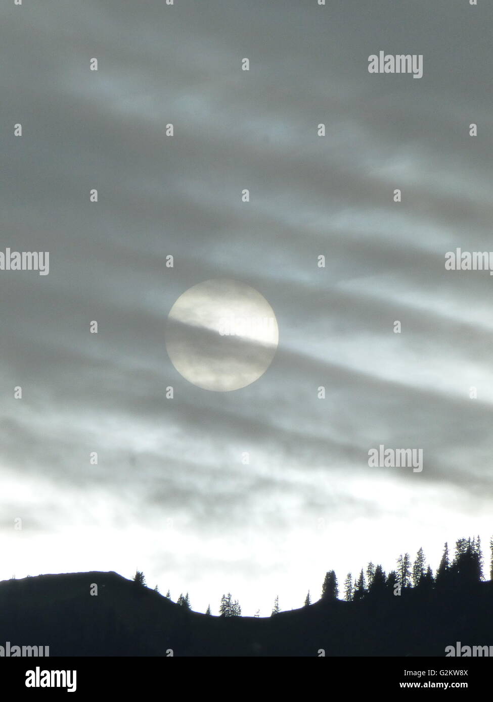 Untergehende Sonne hinter einem Schleier der Wolken mit einem bewaldeten Bergrücken Silhouette im Gegenlicht Stockfoto