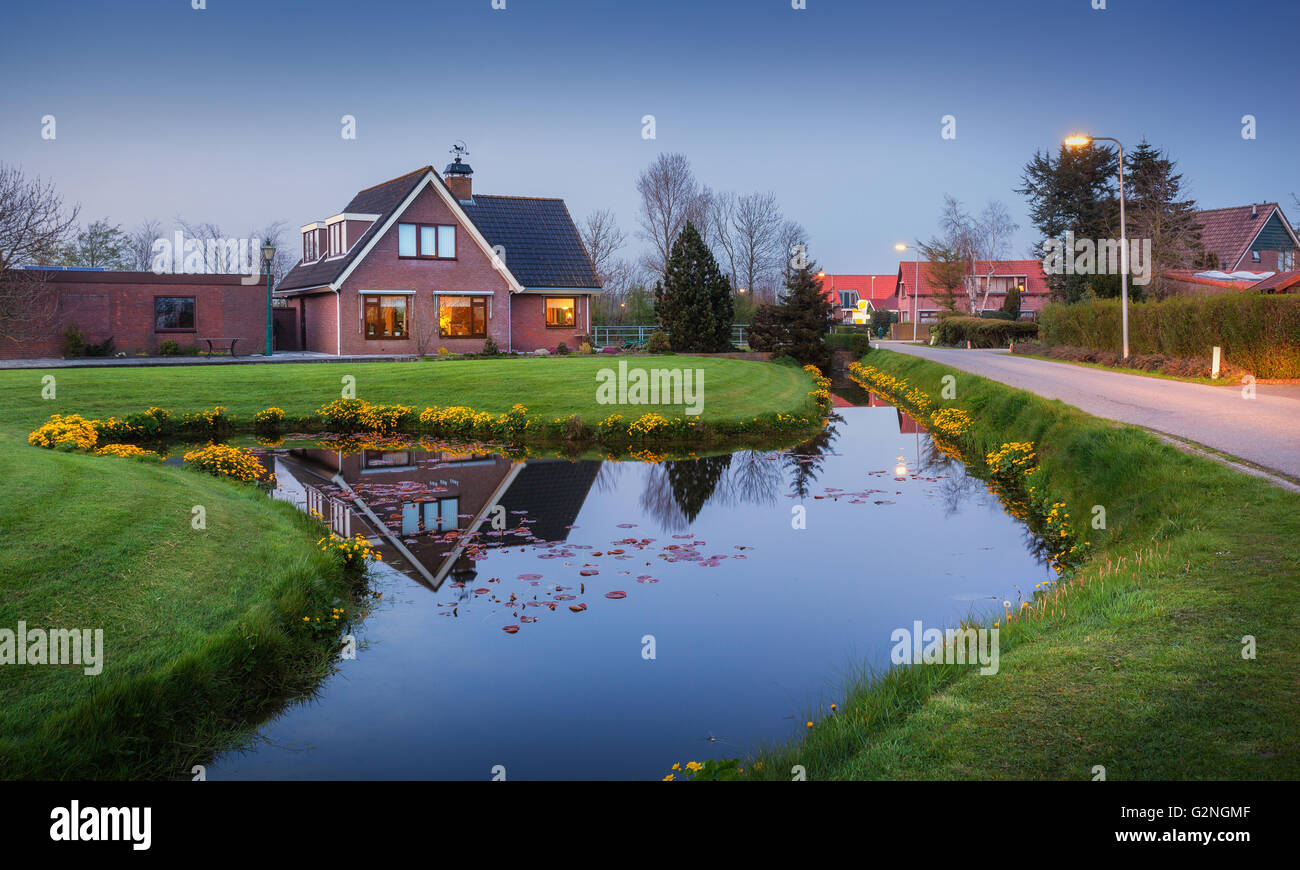 Landschaft im niederländischen Dorf mit schönen Haus spiegelt sich im Wasserkanal, Innenhof mit grünem Rasen, gelben Blüten und Straße w Stockfoto