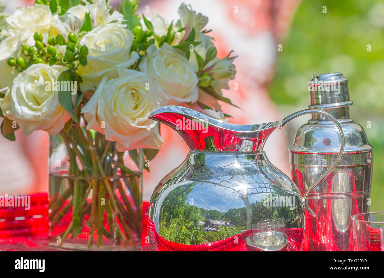 Stillleben mit silbernen Krug und Silber cocktail-Shaker, schneiden frische Rosen in Glasvase mit Wasser am roten Tisch Stockfoto