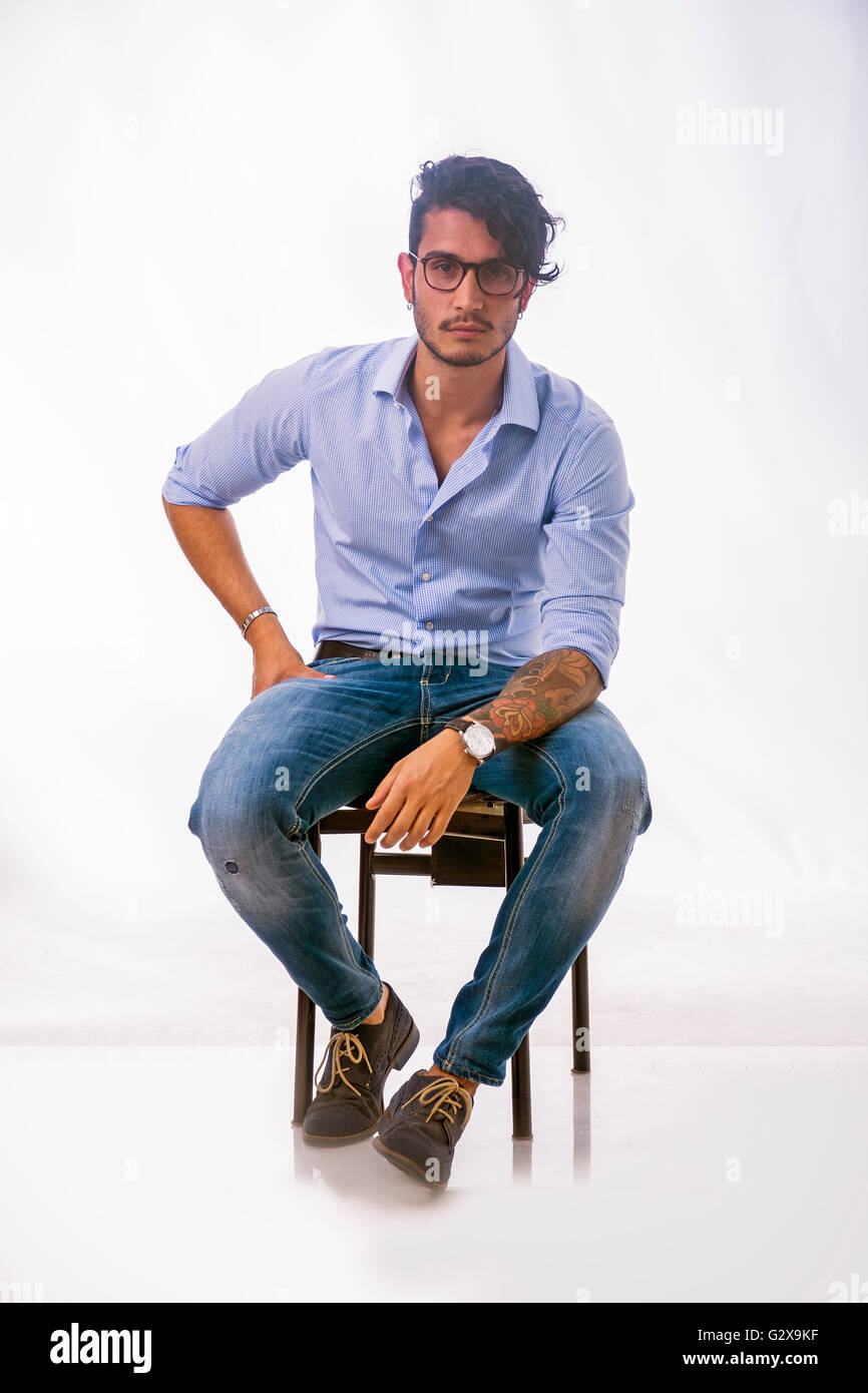 Porträt von Brünette junge Mann in hellblaues Hemd und Jeans, sitzt im Studio gedreht gegen hellen Hintergrund. Foto in voller Länge Stockfoto