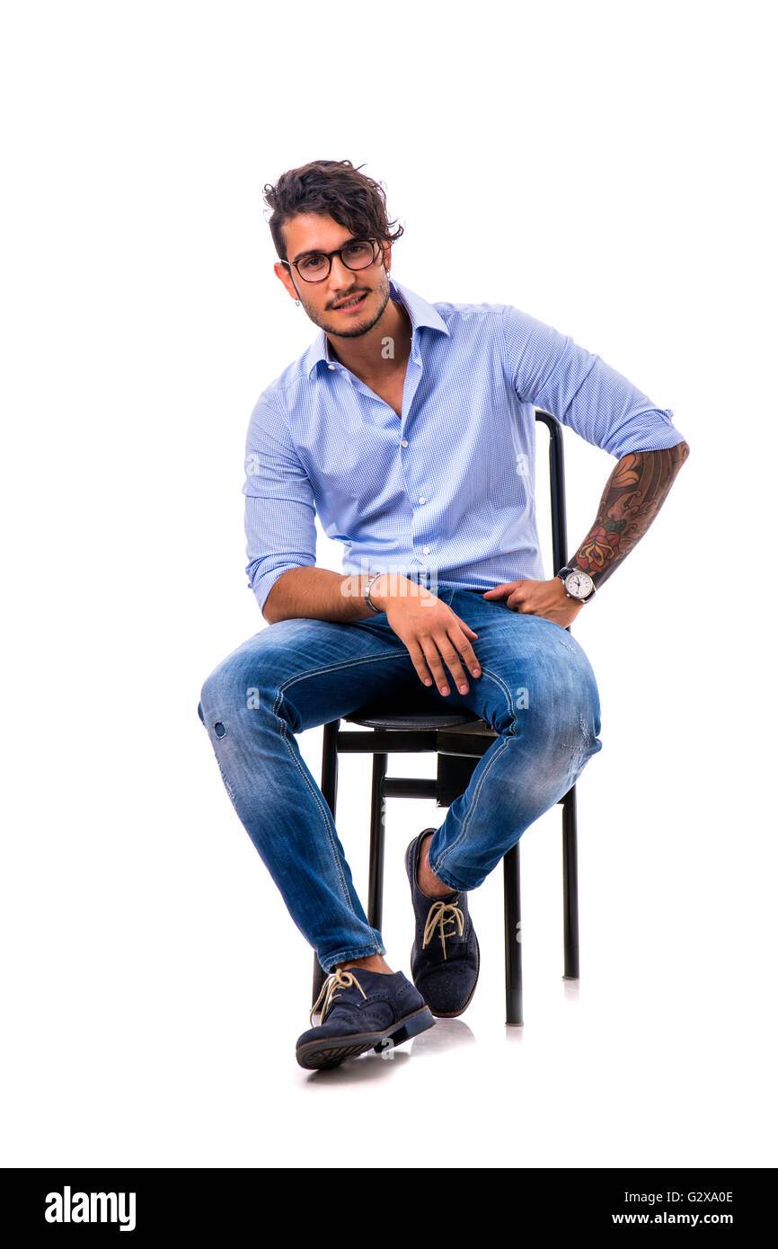 Porträt von Brünette junge Mann hellblaues Hemd und Jeans, vor weißem Hintergrund isoliert sitzen auf Stuhl im Studio gedreht. Stockfoto