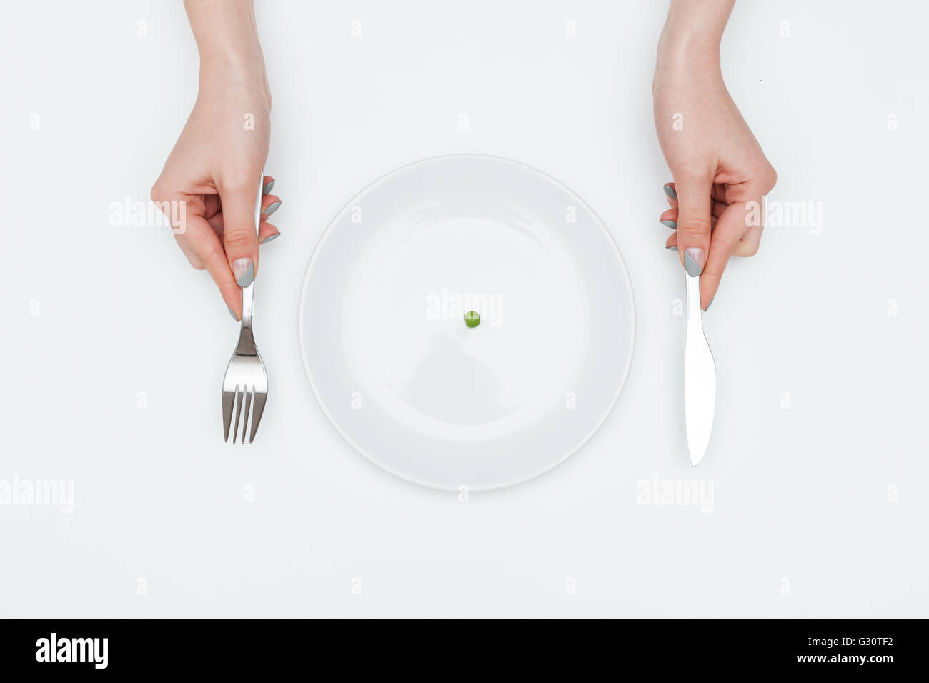 Nahaufnahme der Hände der jungen Frau essen eine kleine grüne Erbse mit Messer und Gabel auf weißem Hintergrund Stockfoto