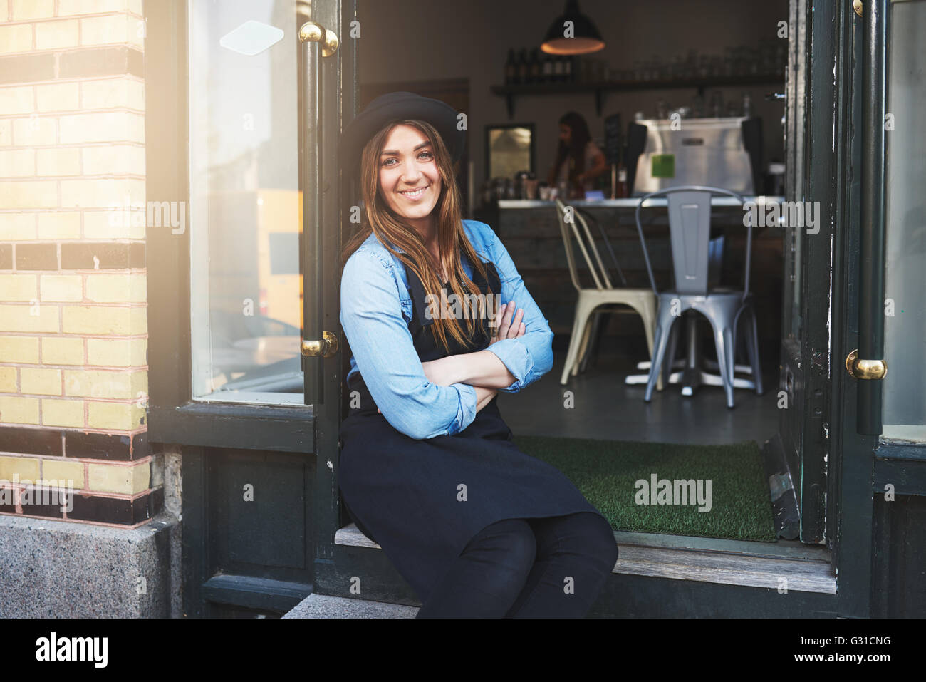 Einzelne nette weibliche Kaffee Hausbesitzer in blauem Hemd, schwarze Schürze und Hut mit verschränkten Armen sitzt in Tür ihres Geschäfts Stockfoto