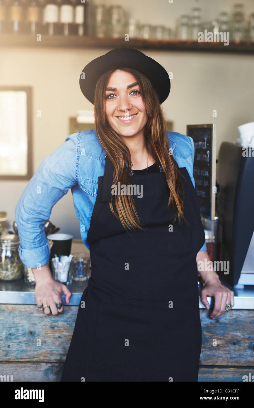 Single weiblich Barista trägt blaue Hemd, schwarze Schürze und Mütze mit fröhlichen Ausdruck gegen Metall und Holz zählen Schiefen Stockfoto