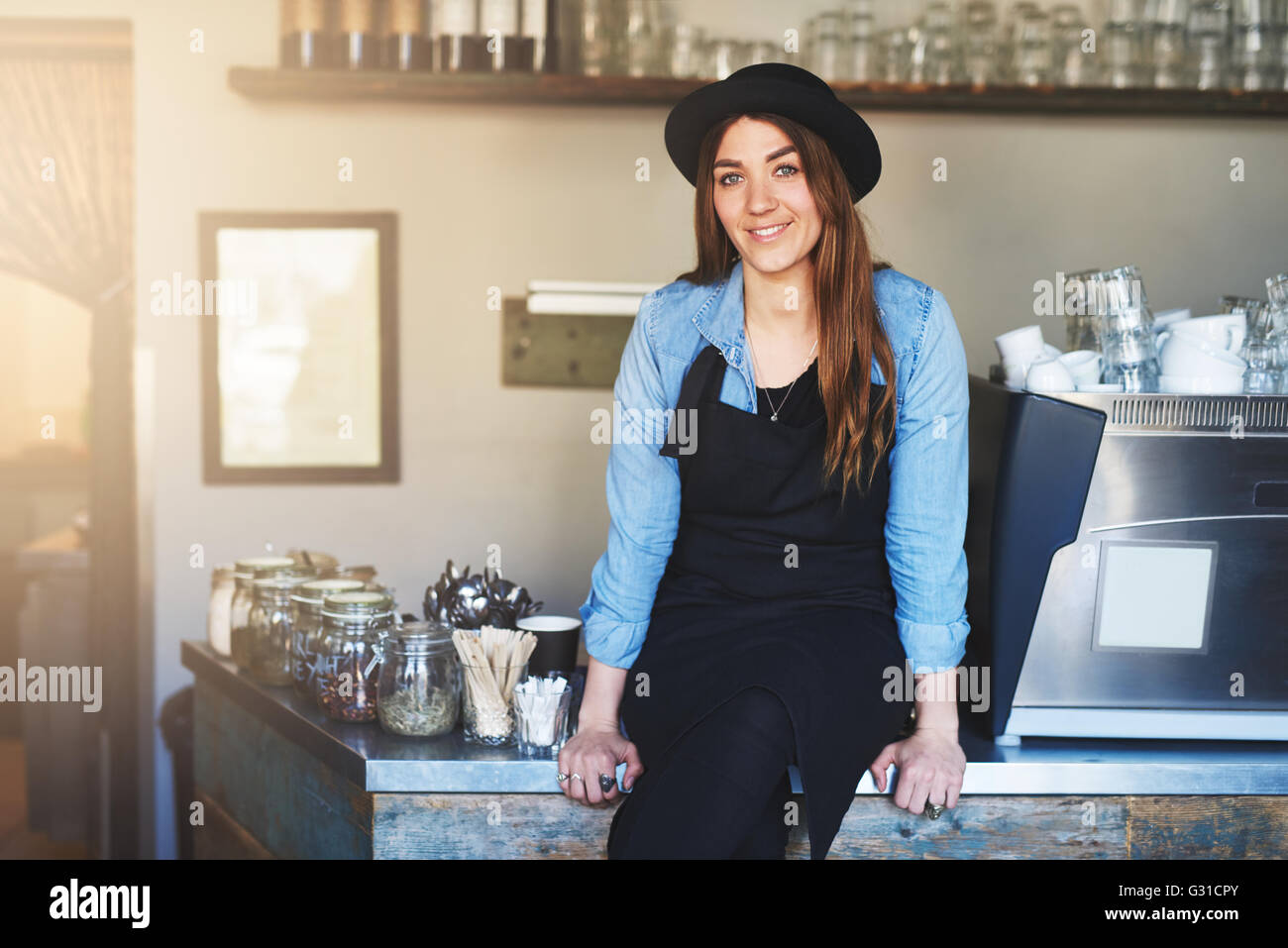 Kaffeehaus-Besitzer sitzt auf Stahl Zähler und lächelt in die Kamera während des Tragens schwarzen Hut und Schürze Stockfoto