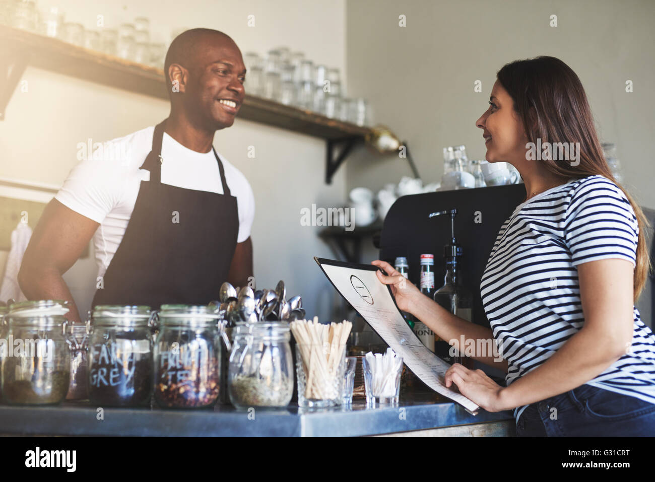 Hübscher Junge schwarze Restaurantbesitzer in Schürze im Gespräch mit fröhlichen Kundin in gestreiften Hemd holding Menü am Schalter Stockfoto