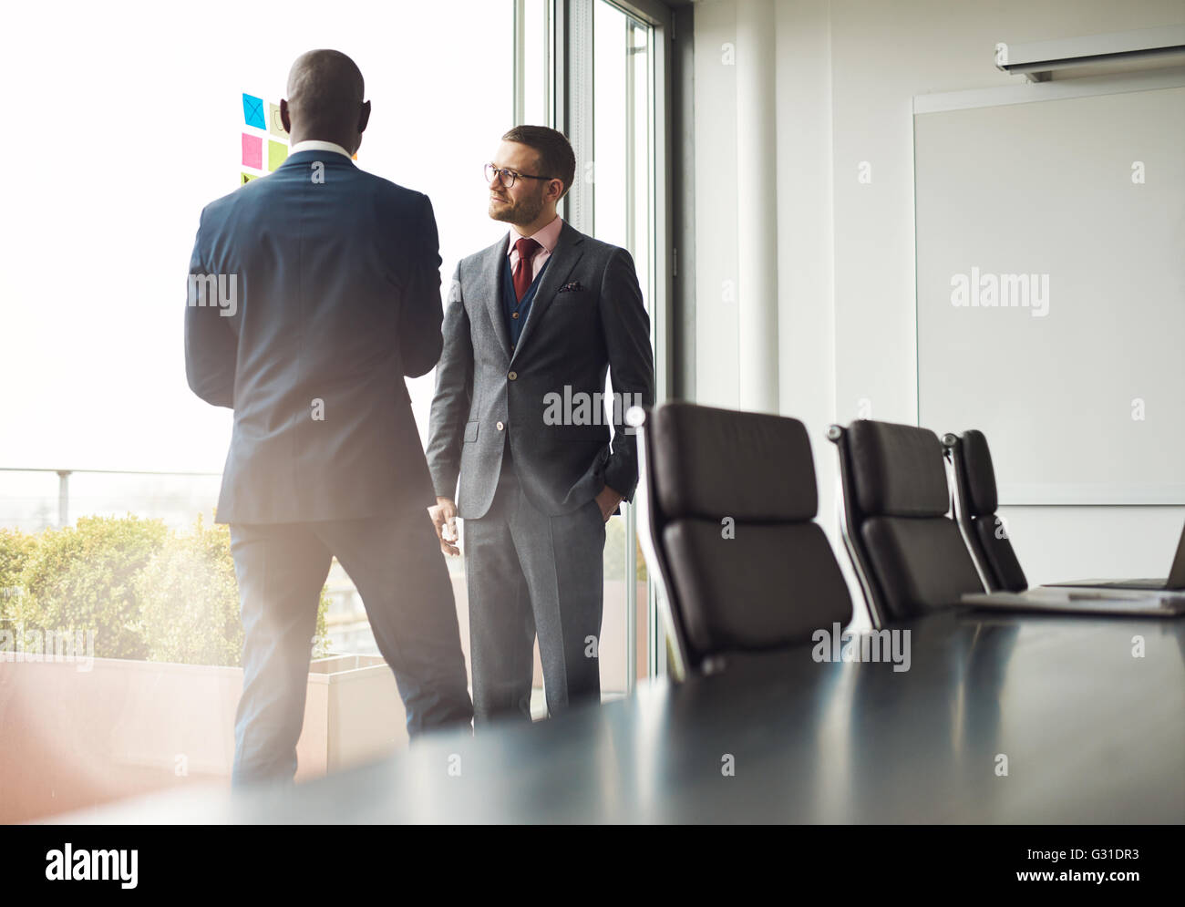 Zwei multirassischen Geschäftsleute stehen zusammen reden vor einem Fenster in einen Konferenzraum, ein Blick auf die Tabelle mit Sun fl Stockfoto