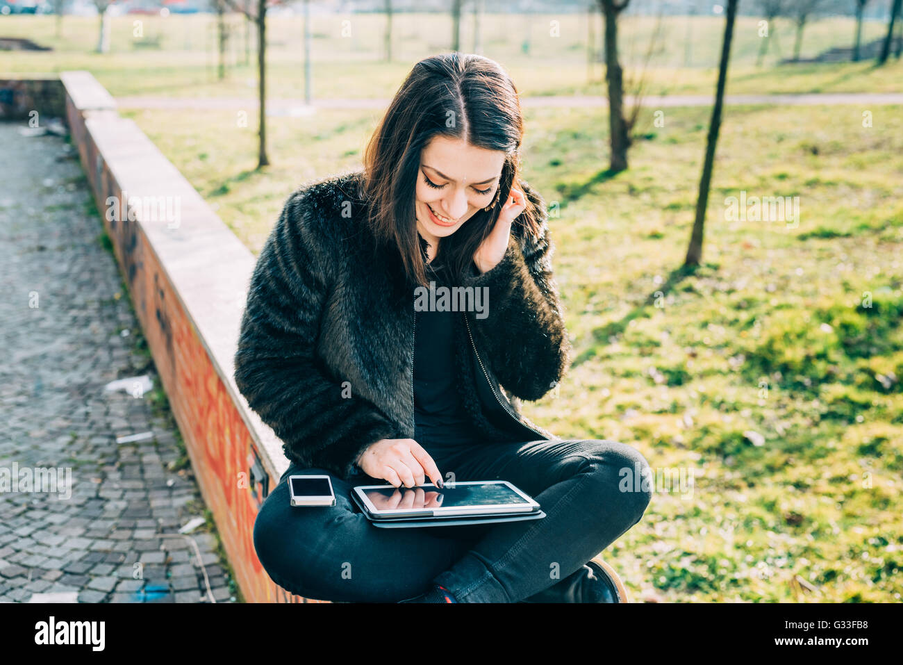 Junge schöne Cacuasian Frau sitzt auf einer kleinen Mauer, die Knie und Smartphone, Tablet über Tippen auf den Touchscreen Lächeln - Multitasking, Technik, Kommunikations-Konzept Stockfoto