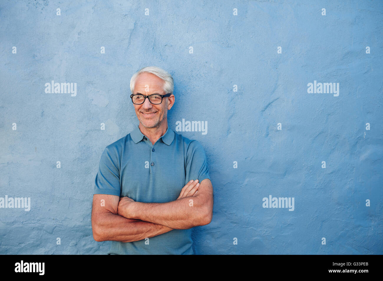 Porträt einer glücklich reifer Mann stehend mit seine Arme verschränkt vor einer blauen Wand. Kaukasischen Mann mit Brille wegsehen ein Stockfoto