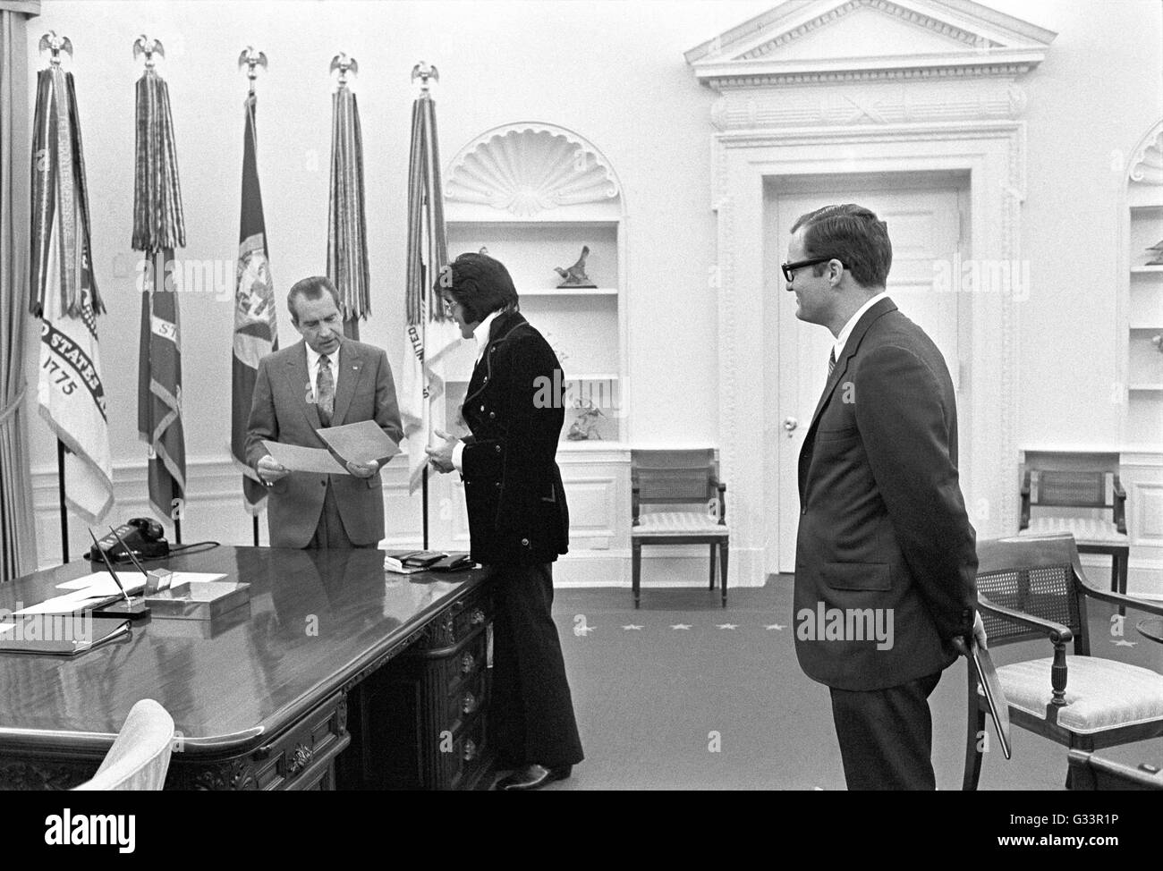 Elvis Presley präsentiert ein signiertes Foto US Präsident Richard M. Nixon als weiße Haus Mitarbeiter sieht Egil Krogh im Oval Office im Weißen Haus 21. Dezember 1970 in Washington, D.C. auf. Das Treffen wurde von Presley, initiiert, Nixon schrieb einen sechs-seitigen Brief mit der Bitte eines Besuchs mit dem Präsidenten und darauf hindeutet, das er ein "Federal Agent-at-Large" im Bureau of Narcotics und gefährlichen Drogen gemacht werden. Stockfoto