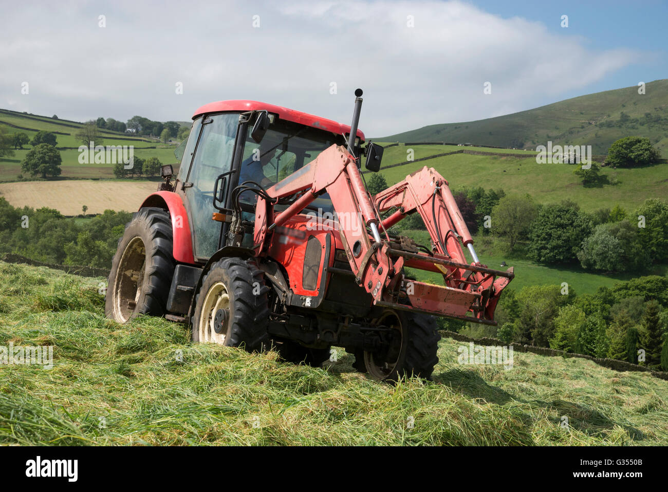 Ein roter Traktor drehen Rasen in einer Sommerwiese Heu in der englischen Landschaft zu trocknen. Stockfoto