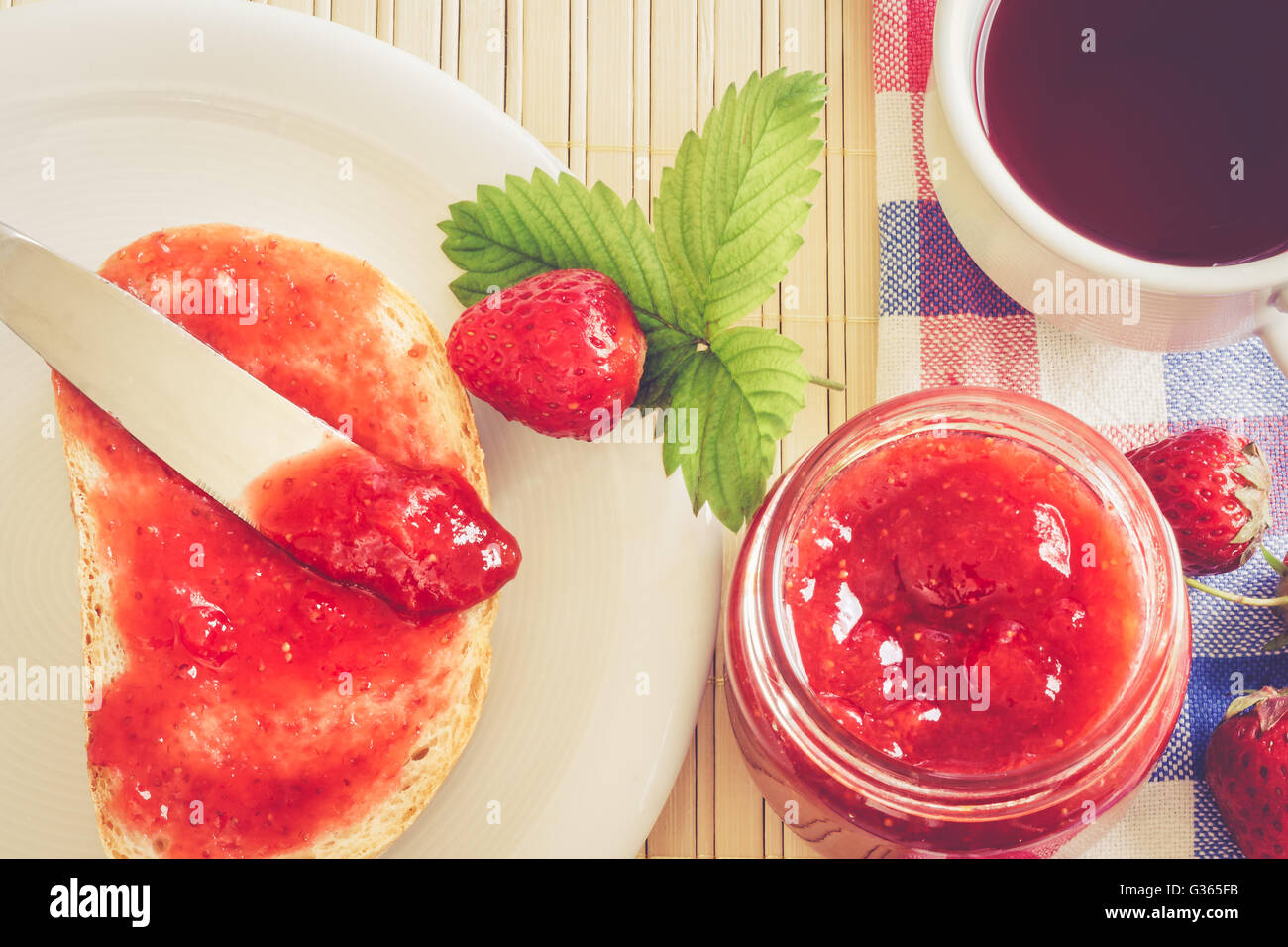 Scheibe Brot mit Glas Erdbeer Marmelade, Tee Teetasse, und frischen Erdbeeren und Blätter neben ihm. Ansicht von oben, Konzept-Bild Stockfoto