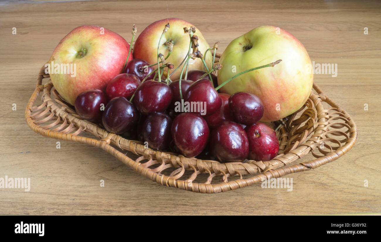 Reifer Apfel und Süßkirschen in geflochtene Platte auf Holztisch Stockfoto