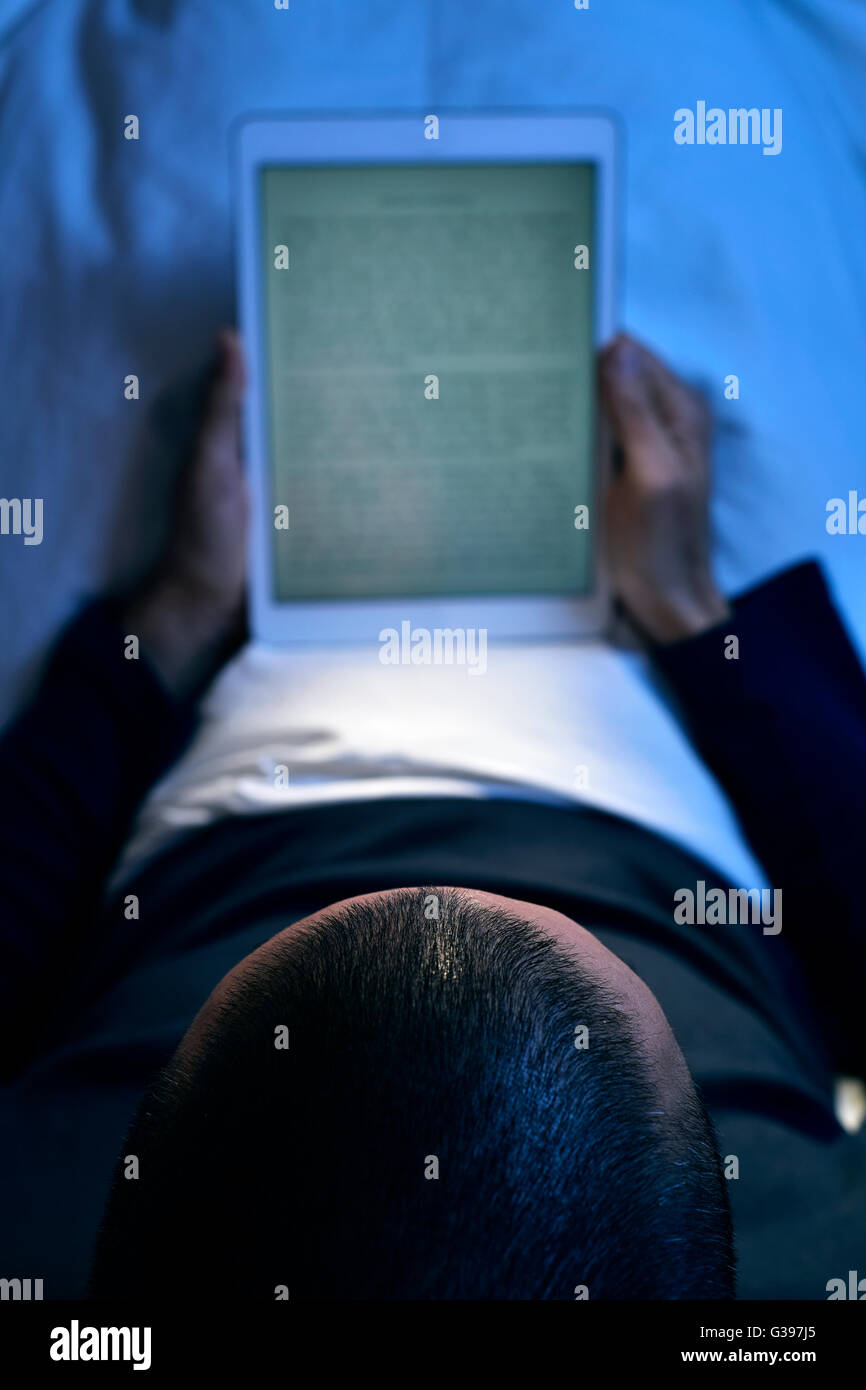 ein junger kaukasischer Mann im Bett liegend liest ein e-Book auf seinem Tablettcomputer in der Nacht Stockfoto