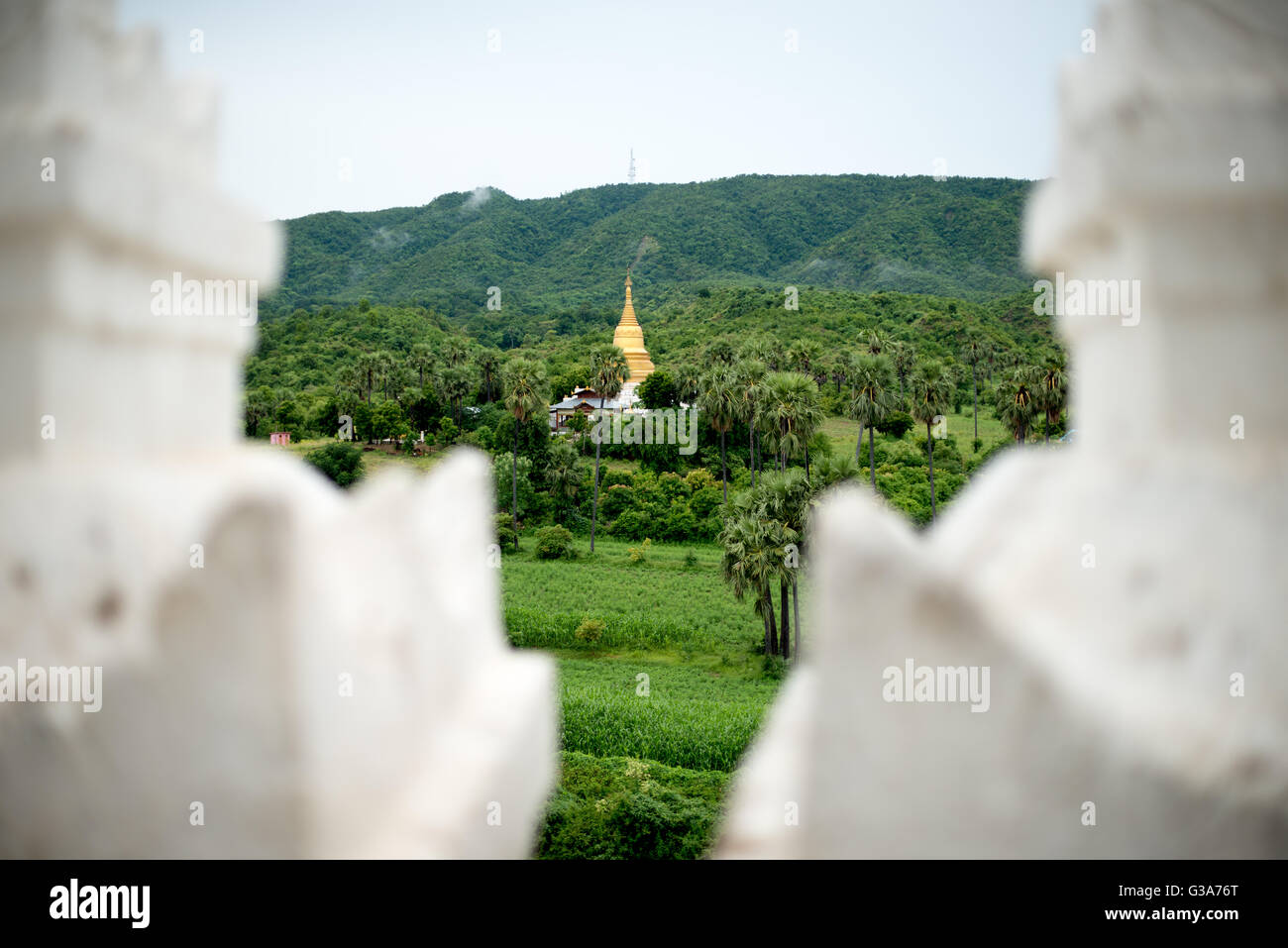 Mingun Pagode Landschaft von Hsinbyume Pagode Myanmar // MINGUN, Myanmar - Eine goldene Stupa erhebt sich zwischen Palmen und üppiger Landschaft, wie von der Hsinbyume Pagode in Mingun aus gesehen. Die Hsinbyume-Pagode, auch bekannt als Myatheindan-Pagode, wurde 1816 von Prinz Bagyidaw als Gedenkstätte für seine erste Frau, Prinzessin Hsinbyume, erbaut. Die weiß gestrichene Pagode stellt den Berg Meru aus der buddhistischen Kosmologie dar und verfügt über sieben wellige Terrassen, die die sieben Bergketten rund um den heiligen Berg symbolisieren. Mingun liegt am westlichen Ufer des Ayeyarwady River, etwa 11 km nördlich von Stockfoto