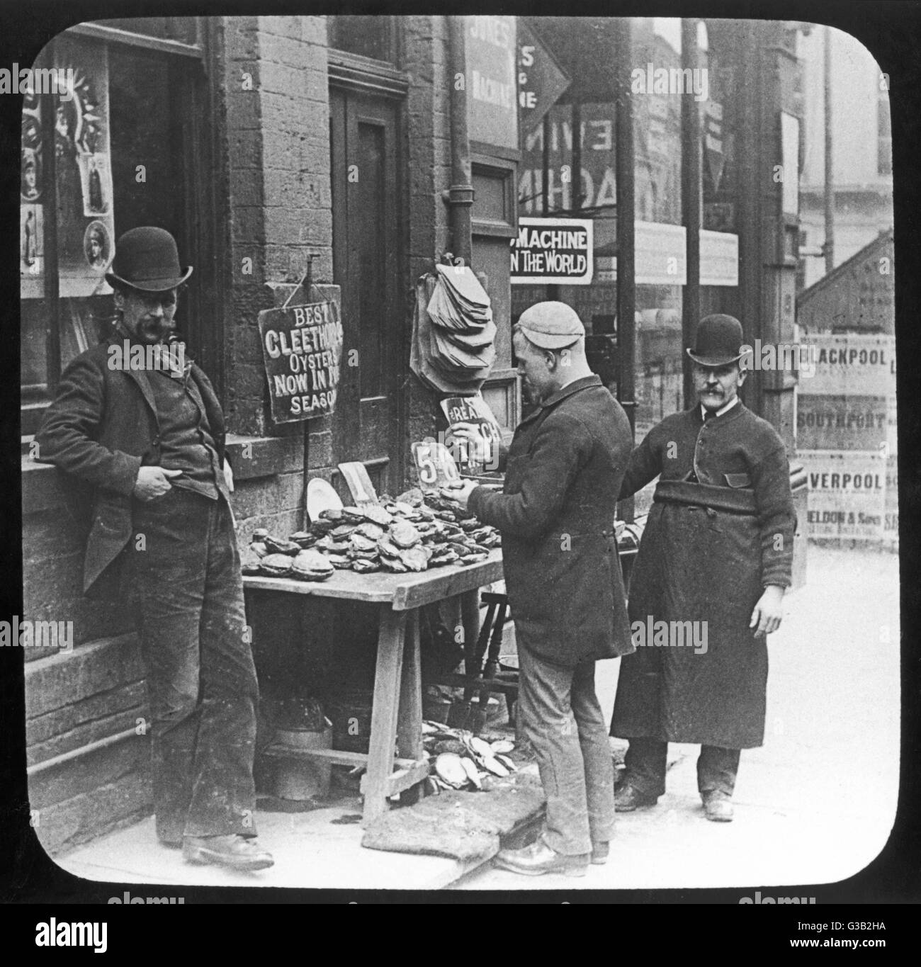 Ein Bowler hüten Auster Verkäufer sieht, während ein Arbeiter mit seiner Mütze an der Seite seines Kopfes selbst Cleethorpes Austern hilft.     Datum: um 1890 Stockfoto