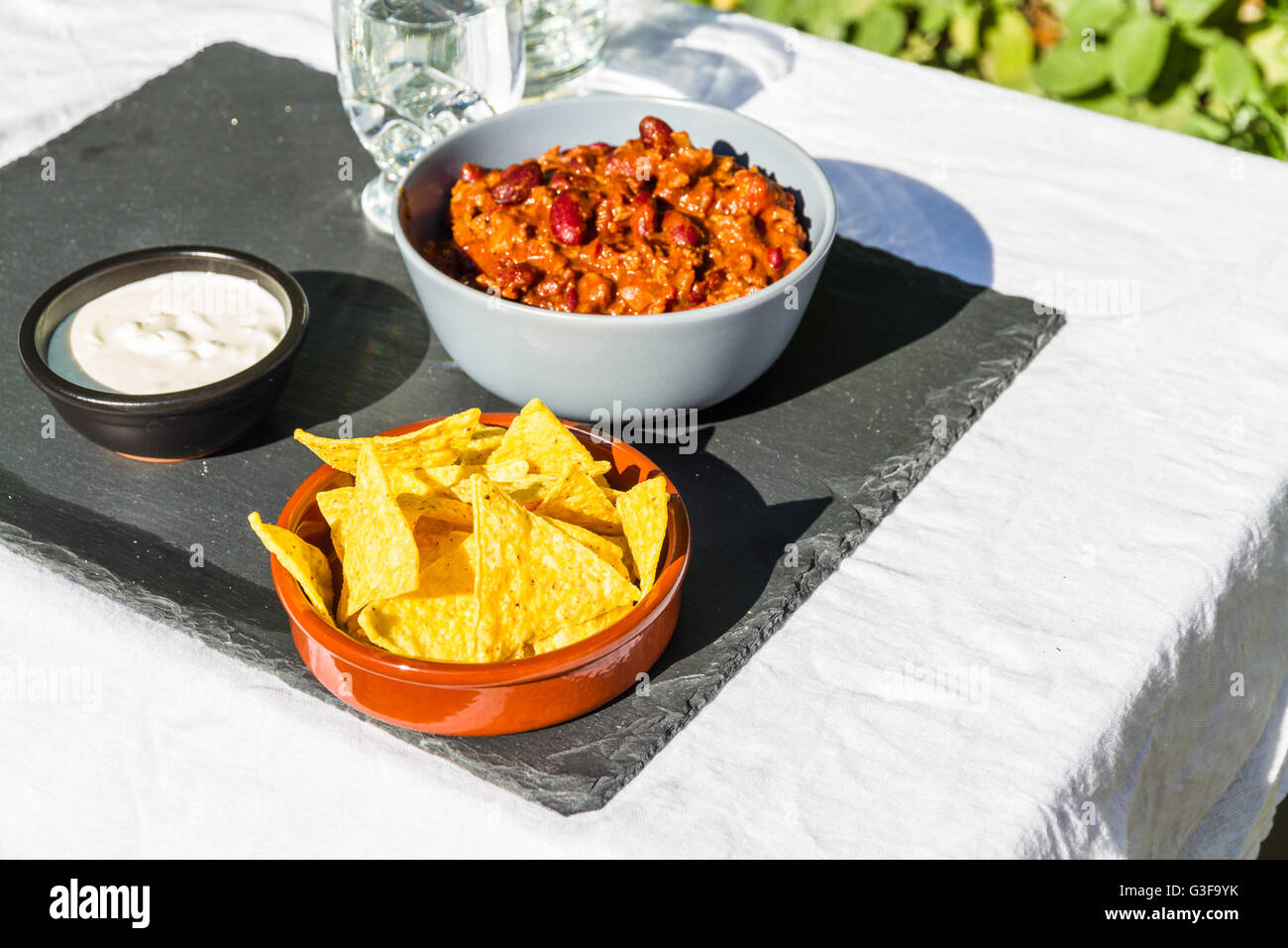 Hausgemachte Chili Con Carne in orange Keramik Schüssel und Tortilla-Chips und saure Sahne mit Krug und Glas Eistee Wat auf der Seite Stockfoto