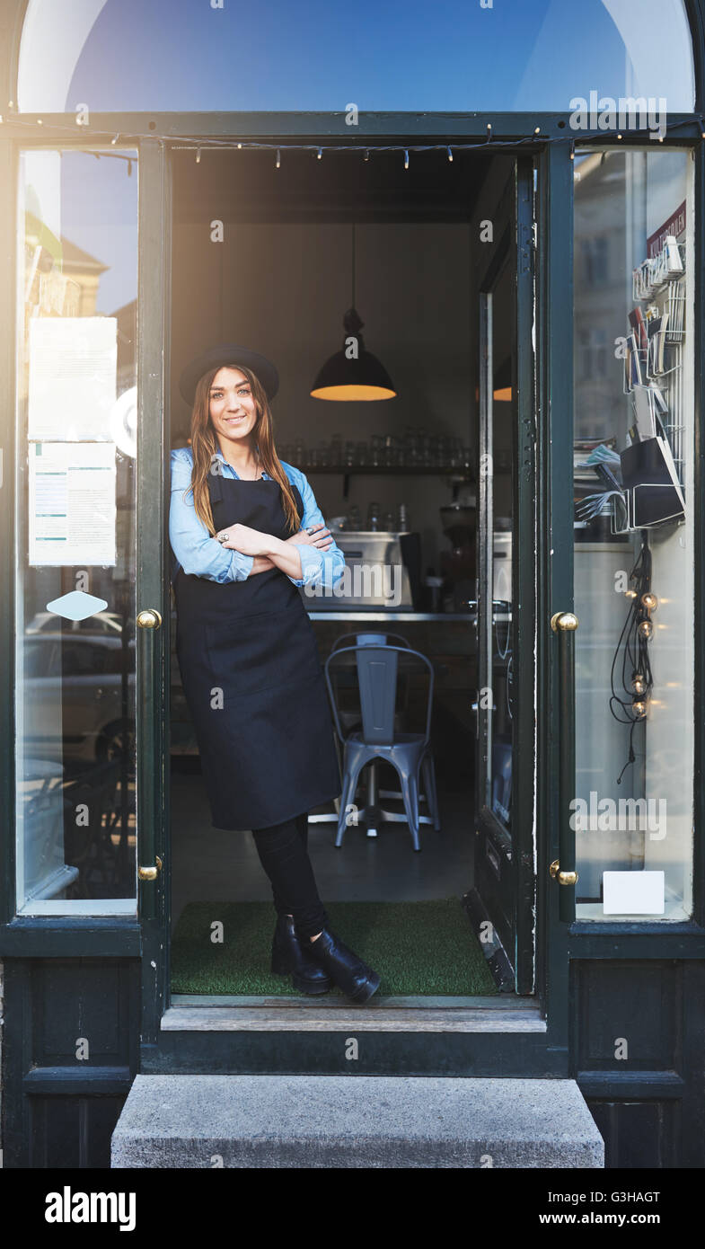 Einzelne nette weibliche Barista in Schürze und Hut mit verschränkten Armen gelehnt Eingang im café Stockfoto