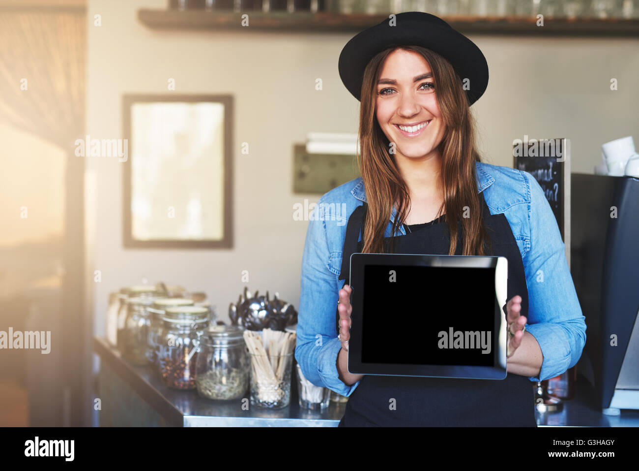 Einzelne niedlich Kaffee Haus Arbeiter in Hut und Schürze mit fröhlichen Ausdruck leere Tablet-Computer in der Nähe von Zähler hält Stockfoto
