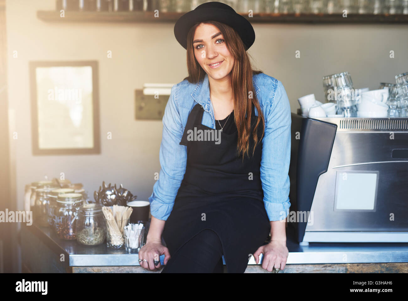 Weibliche Barista lächelnd sitzt auf Café-Zähler mit einem schwarzen Hut und Schürze über einem Hemd Langarm blau Jean neben Reihen von gl Stockfoto