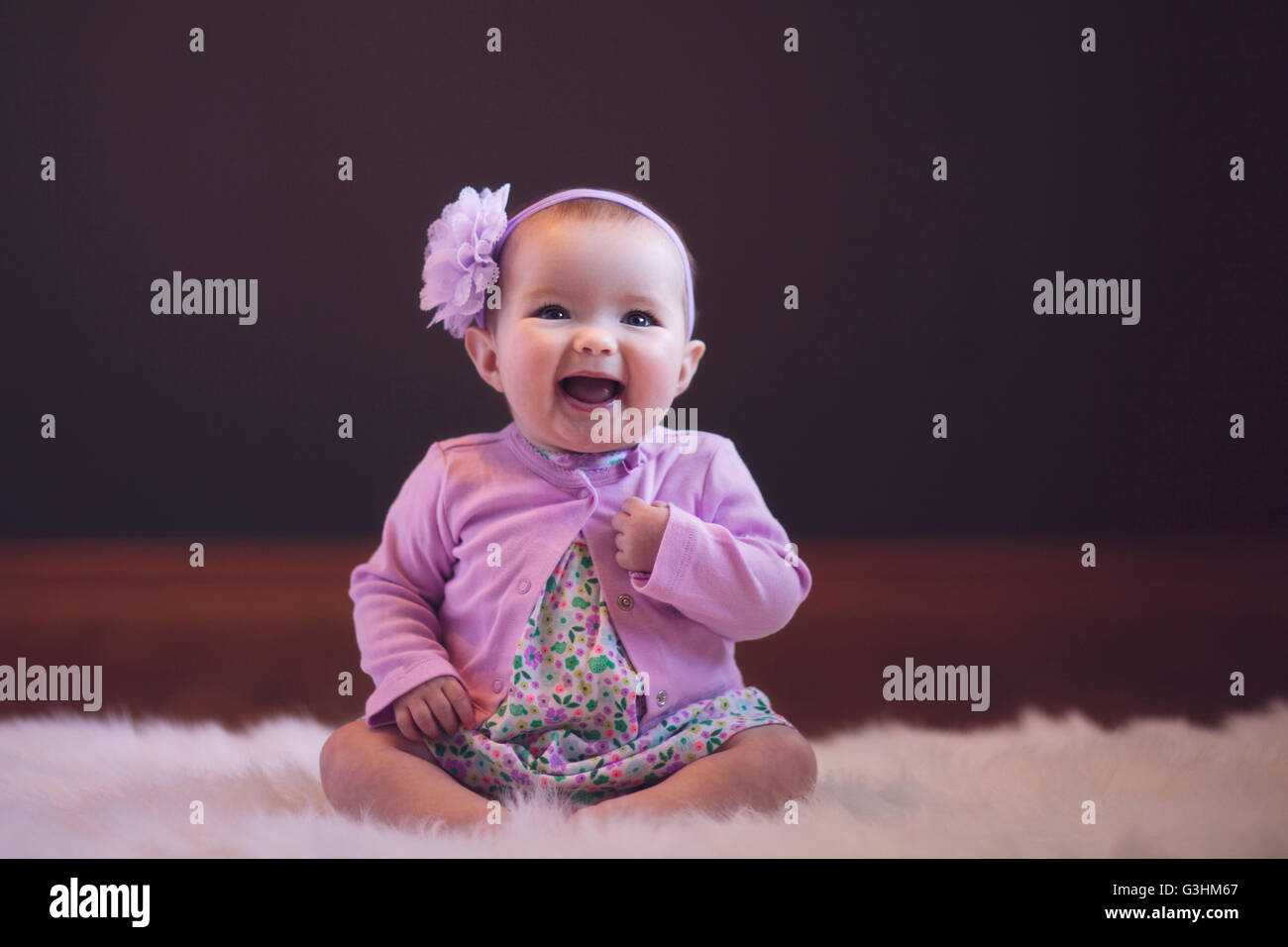 Babymädchen sitzen auf Teppich tragen Blütenkopf Band Blick auf Kamera Mund öffnen Sie lächelnd Stockfoto